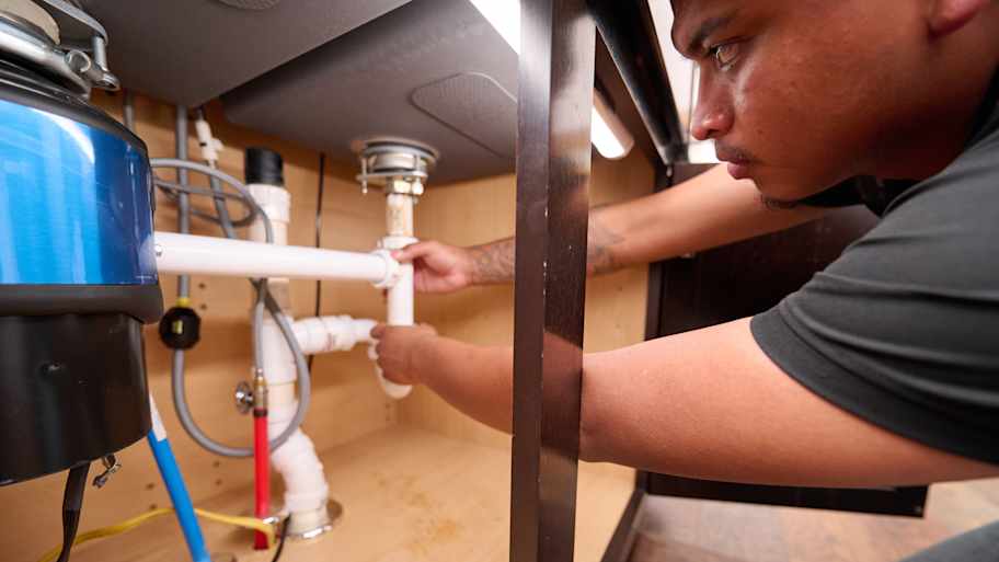 Plumber installing PVC pipes under a kitchen sink, connecting drainage system and garbage disposal in a cabinet space.