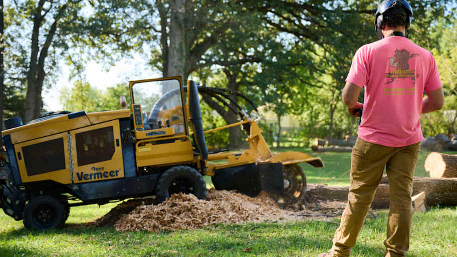 Tree trimmer in safety helmet observes a yellow Vermeer stump grinder mulching a tree stump in a sunny, grassy area.