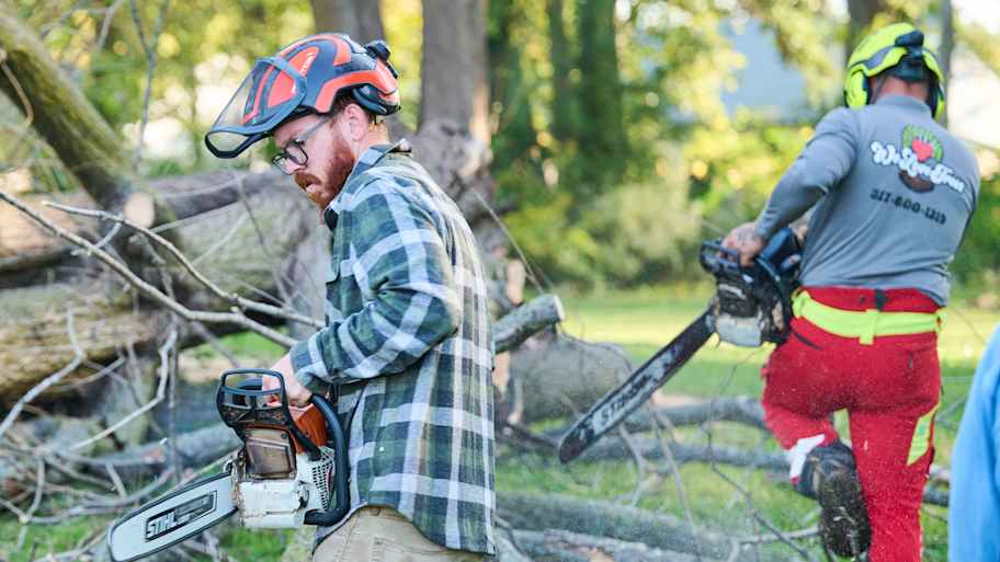 Two workers with chainsaws clear fallen tree branches in a grassy area surrounded by trees one wearing flannel and the other in safety gear.