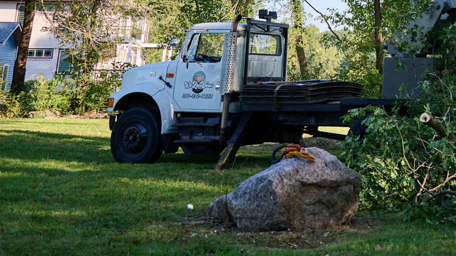 White flatbed truck with company logo is parked on grass near a large boulder and tree debris in a residential area.