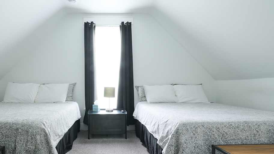 A bright bedroom with two large beds, dark curtains, a central nightstand, and a ceiling fan, creating a cozy ambiance.