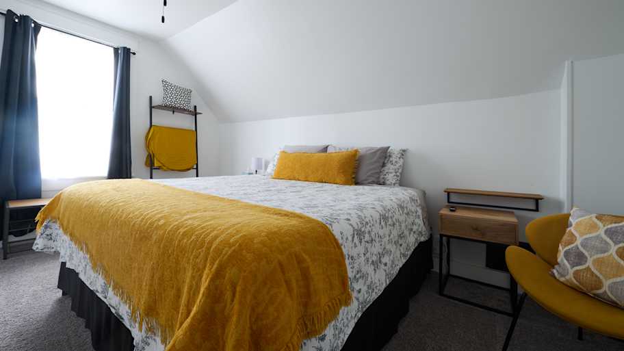 A cozy bedroom with a sloped ceiling, featuring a bed with floral bedding and a mustard yellow throw, and modern furnishings.