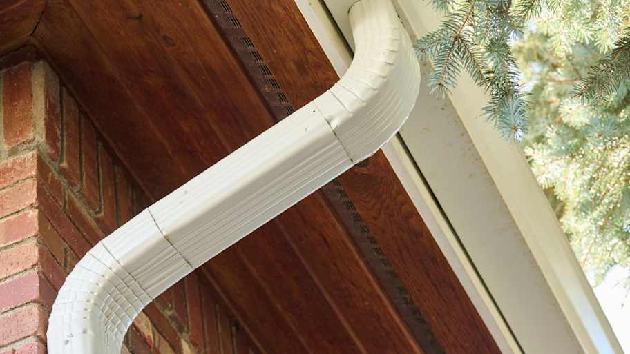 White downspout with curved elbow section attached to brick wall, directing water runoff from roof to ground level.
