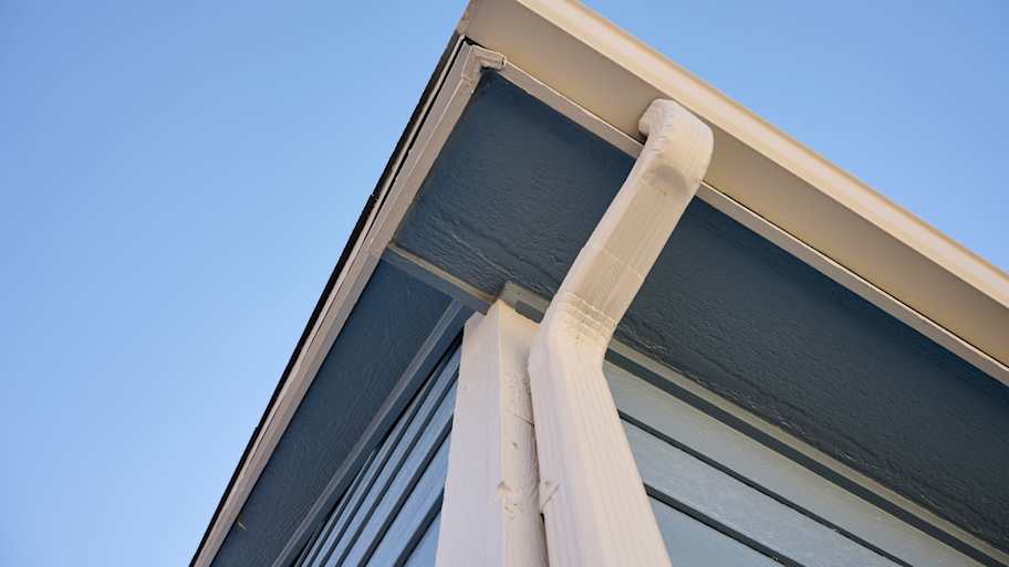 White downspout connected to seamless gutter on a blue-painted home exterior, channeling rainwater away effectively.