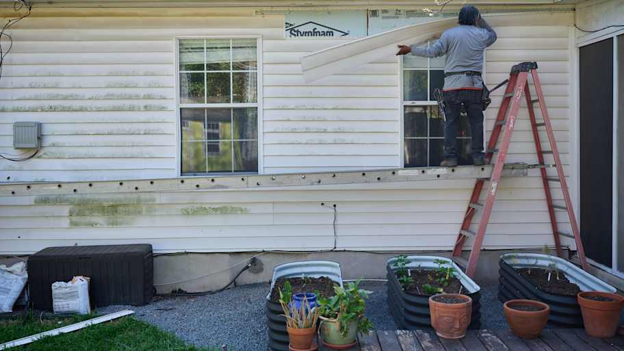 Worker replacing exterior vinyl siding on a residential home using a ladder and scaffold, with garden planters below