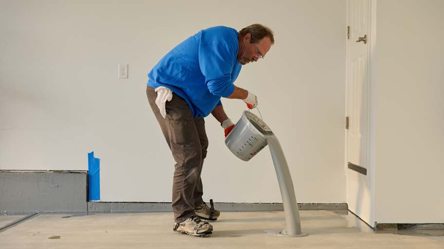 Worker pouring gray epoxy coating onto a concrete floor, preparing the surface for a smooth, durable finish.