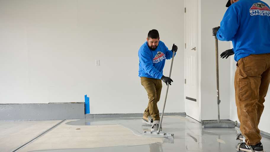 Two workers in blue shirts spreading gray epoxy coating on a garage floor using squeegees for a smooth, even finish.