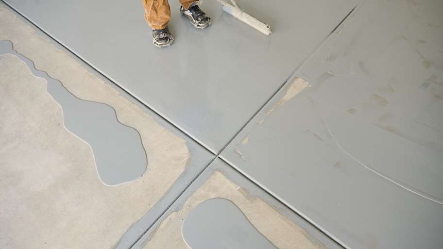 Worker applying gray epoxy coating to concrete floor, with patches of wet epoxy and visible roller tool in use