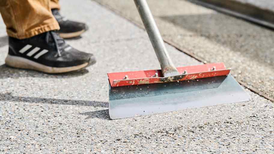 Worker using a red-edged floor scraper to smooth and level concrete surface with a textured, pebble-finish coating