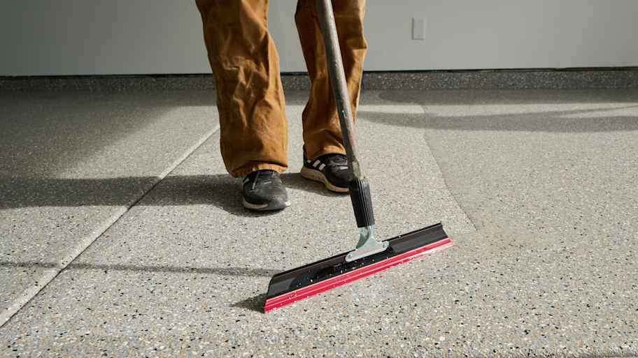 Worker using a red-edged squeegee to spread epoxy coating over a speckled garage floor for a smooth, uniform finish.