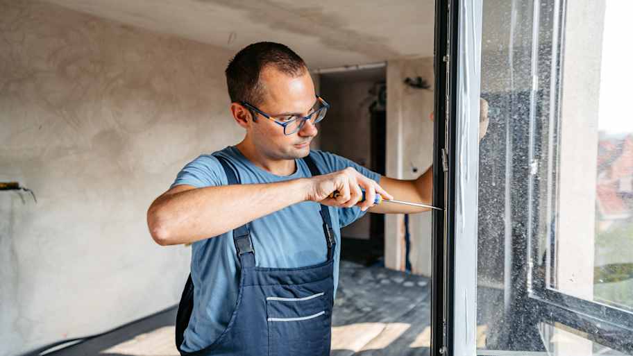 Repairman in overalls screwing the windows in an empty apartment under renovation.