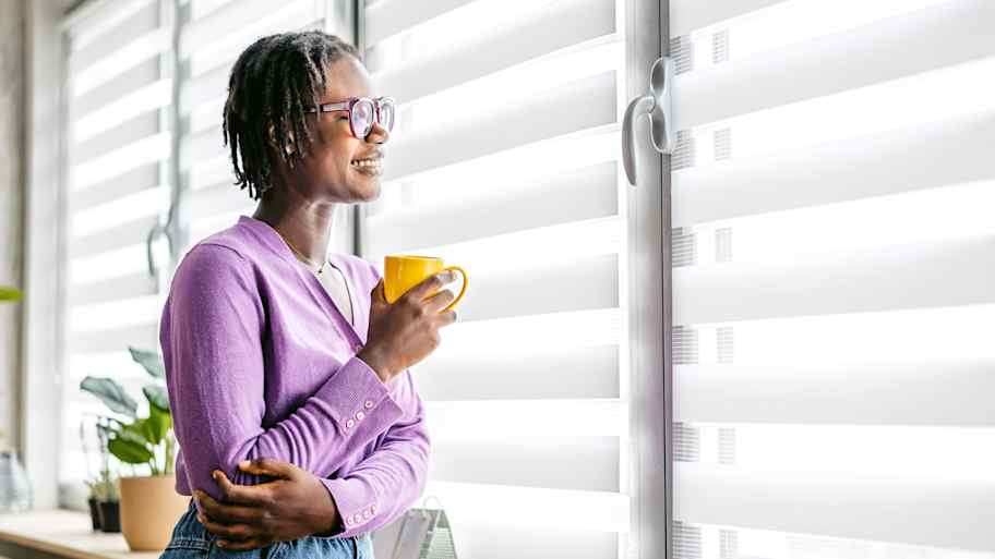 Woman drinking coffee and looking out a window with blinds  