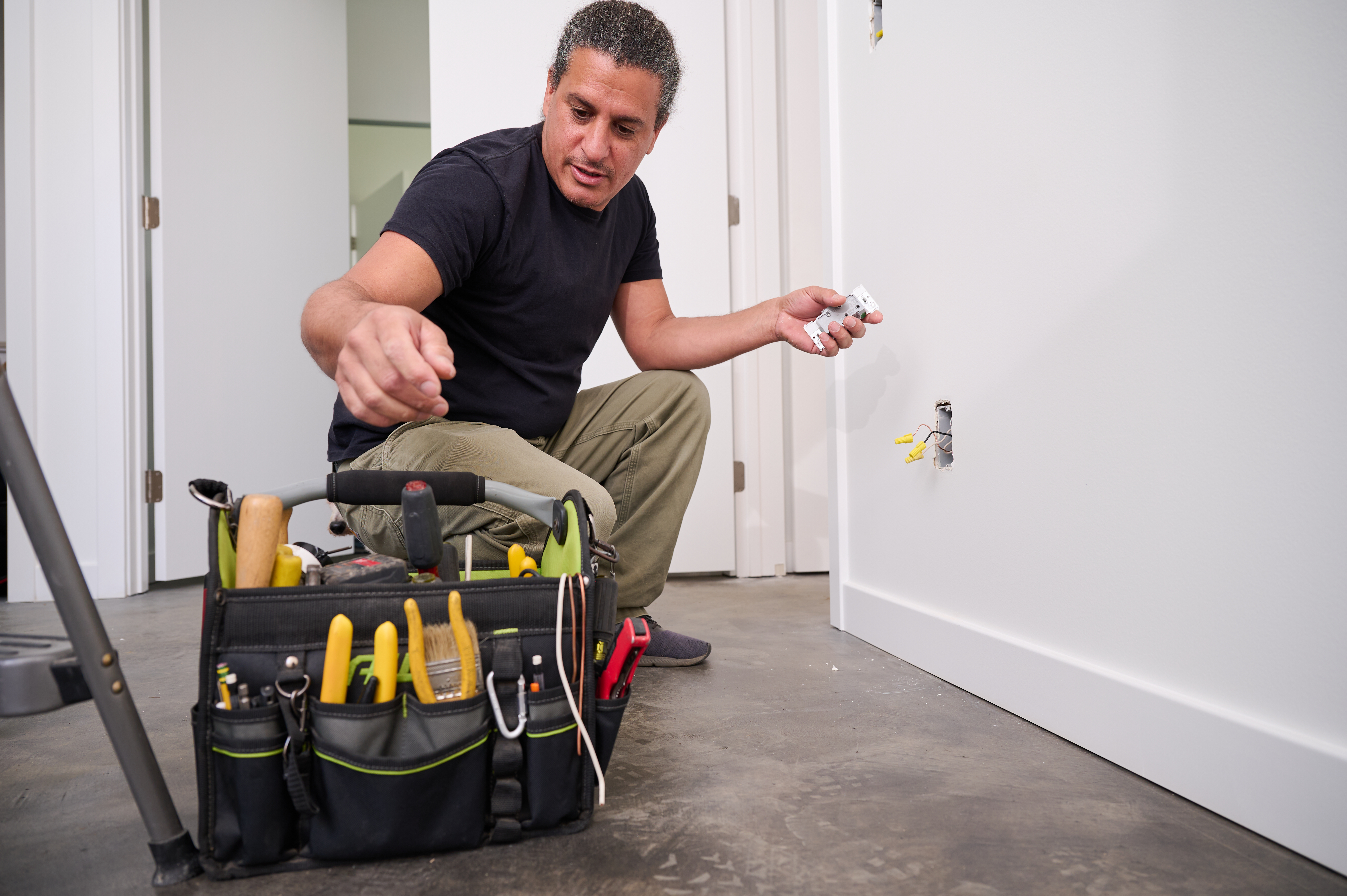 A technician kneels to install an electrical outlet, holding the outlet in one hand while selecting tools from a nearby tool bag for the job.