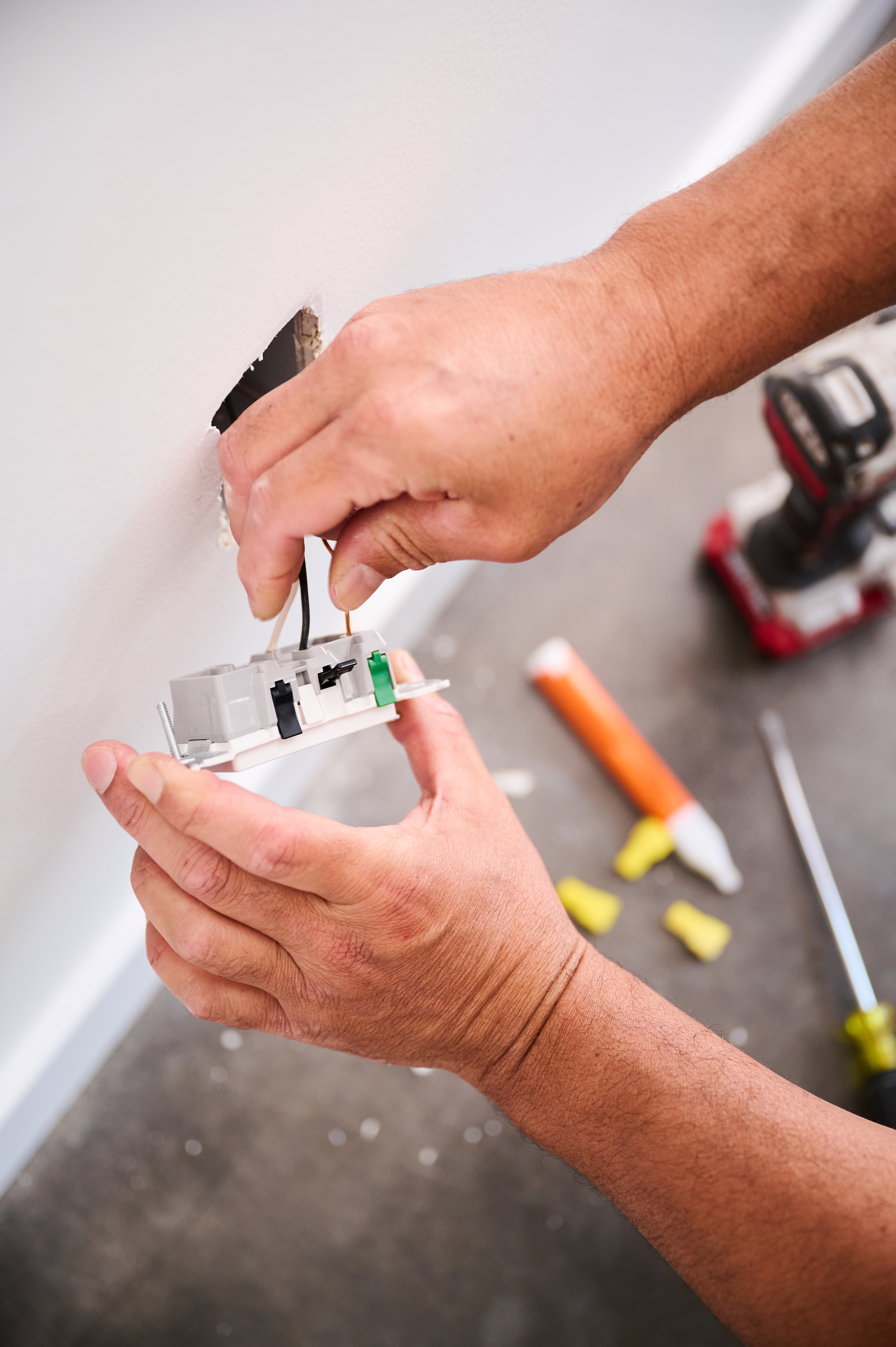 Close-up of a technician's hands connecting black and copper wires to a wall socket, with a power drill and other tools in the background.