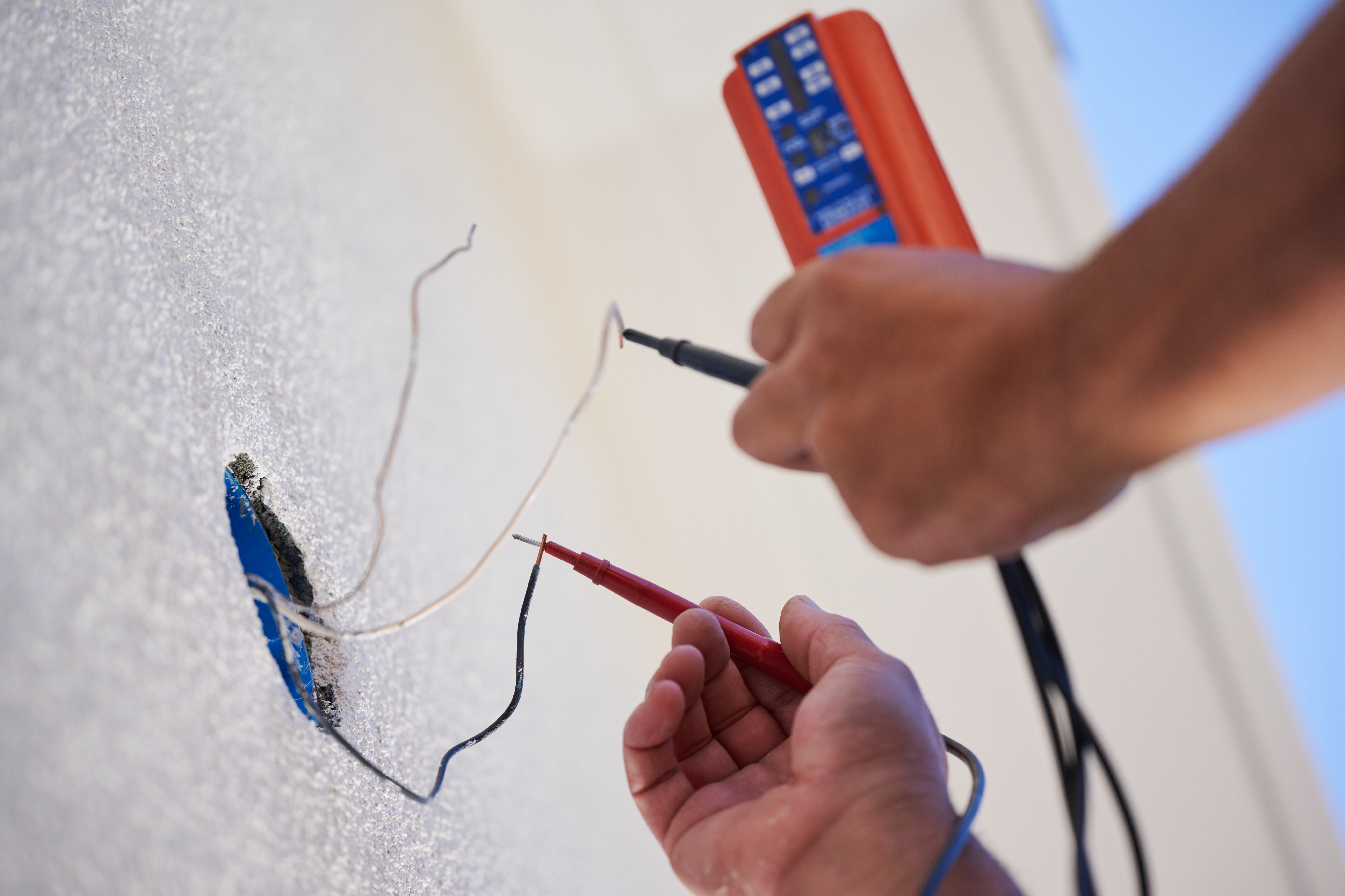 A close-up of a technician testing exposed wires with a multimeter to ensure proper electrical connectivity on a white wall installation.