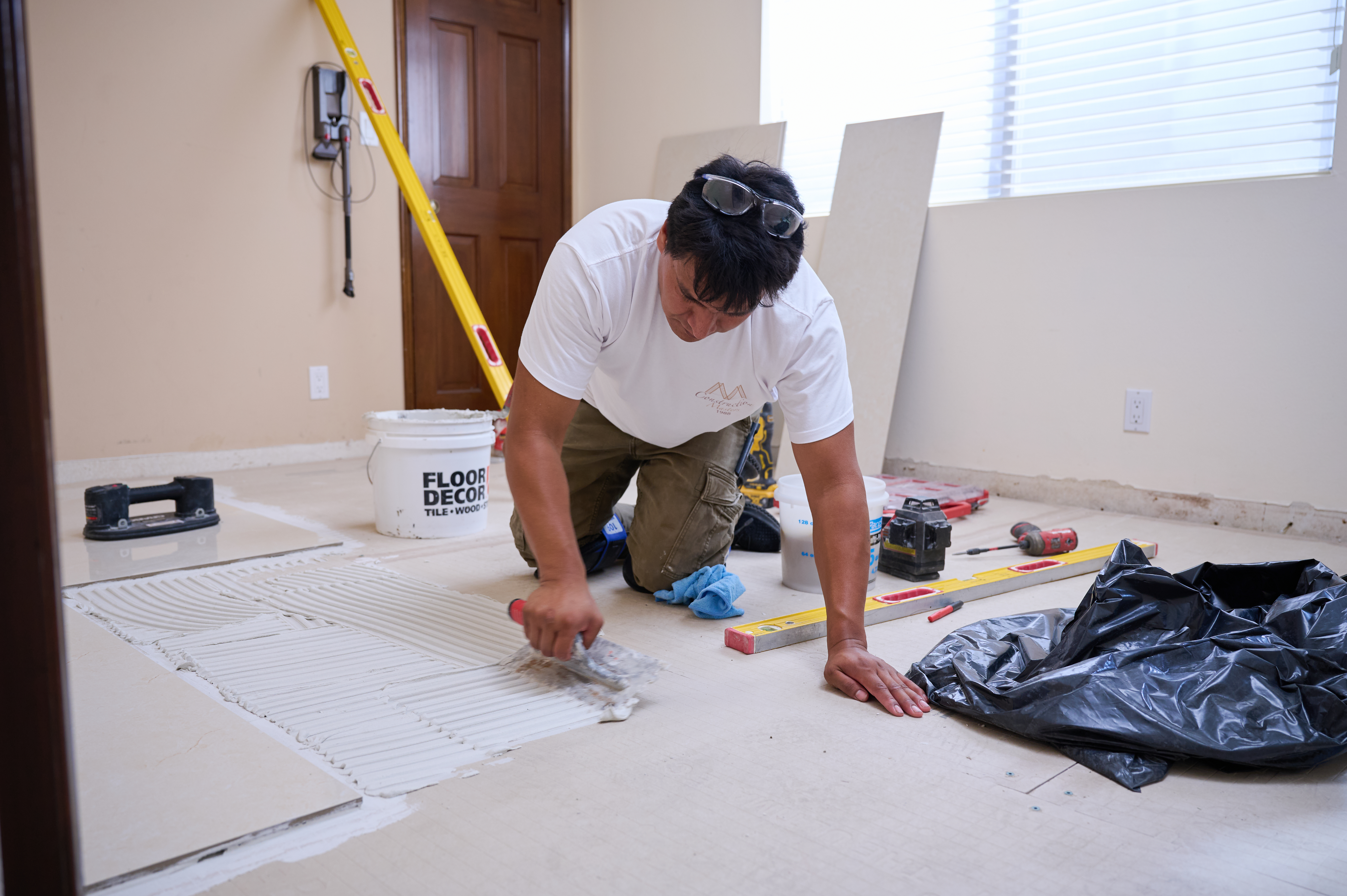 A worker applies tile adhesive to the floor with a trowel, preparing the surface for tile installation in a home renovation project.