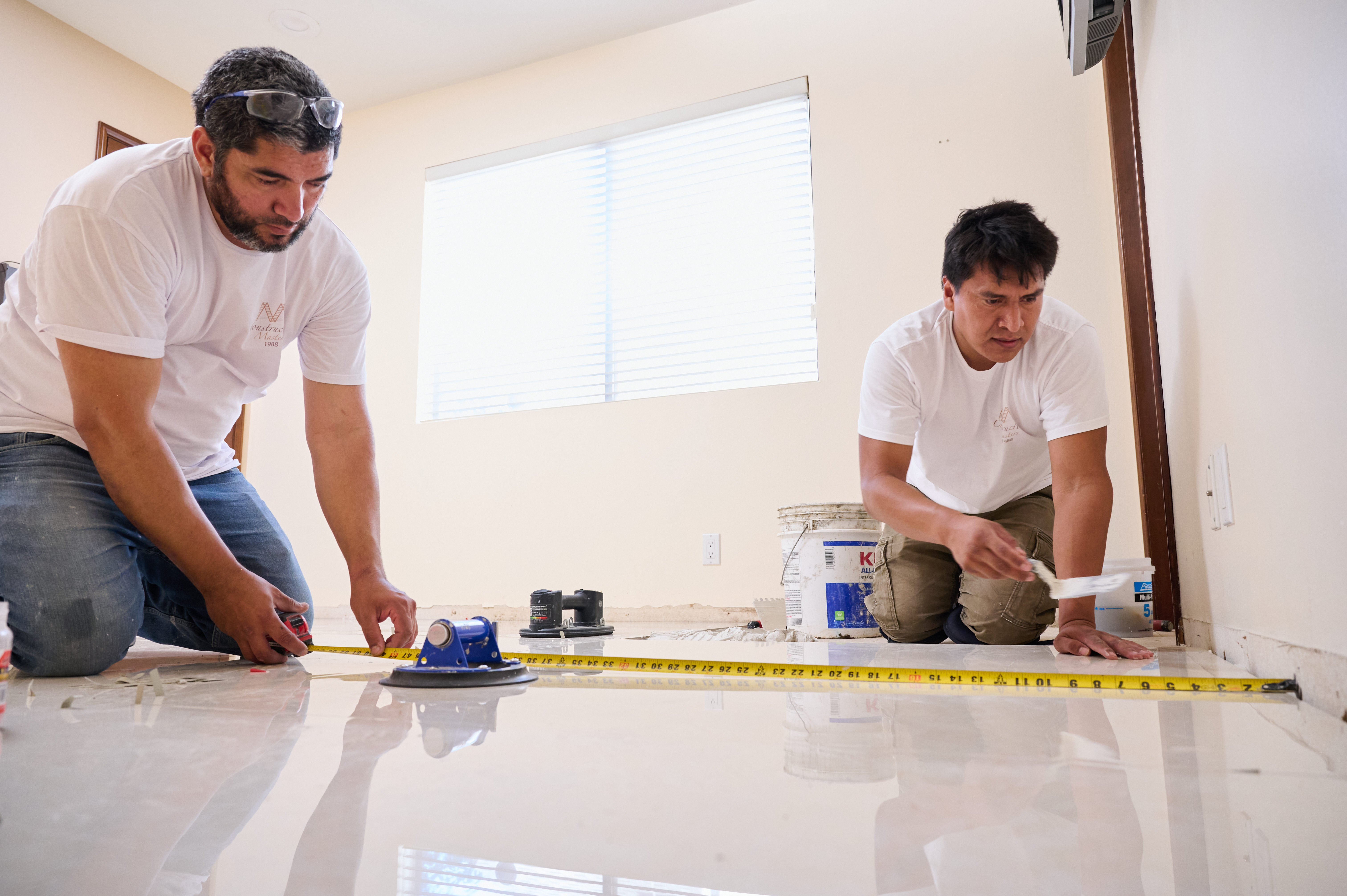 Two workers measure and prepare the floor for tile installation, using a tape measure and tools for accurate placement.