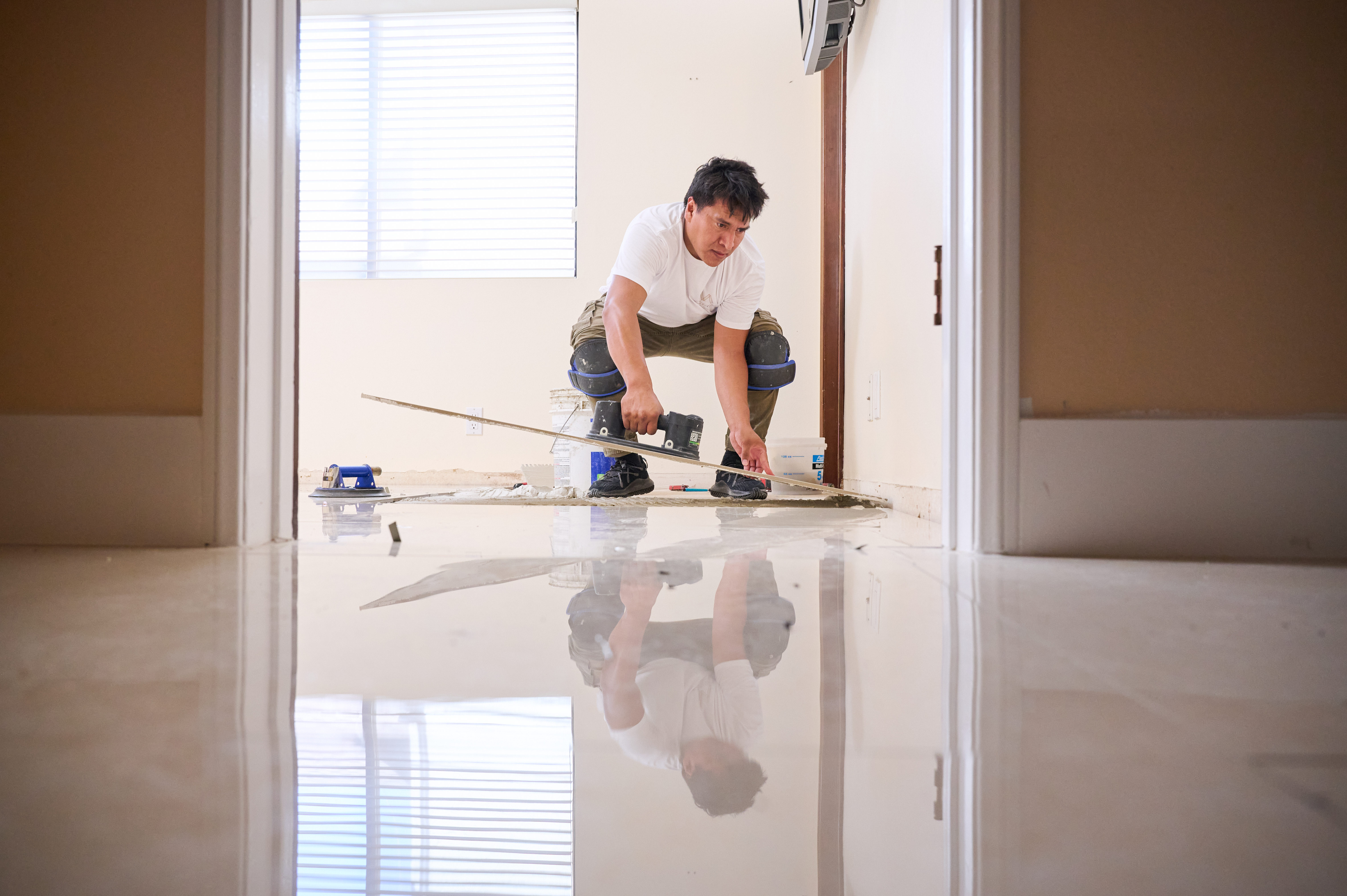 A worker in knee pads carefully aligns a tile during floor installation, with a polished floor reflecting his focused posture.