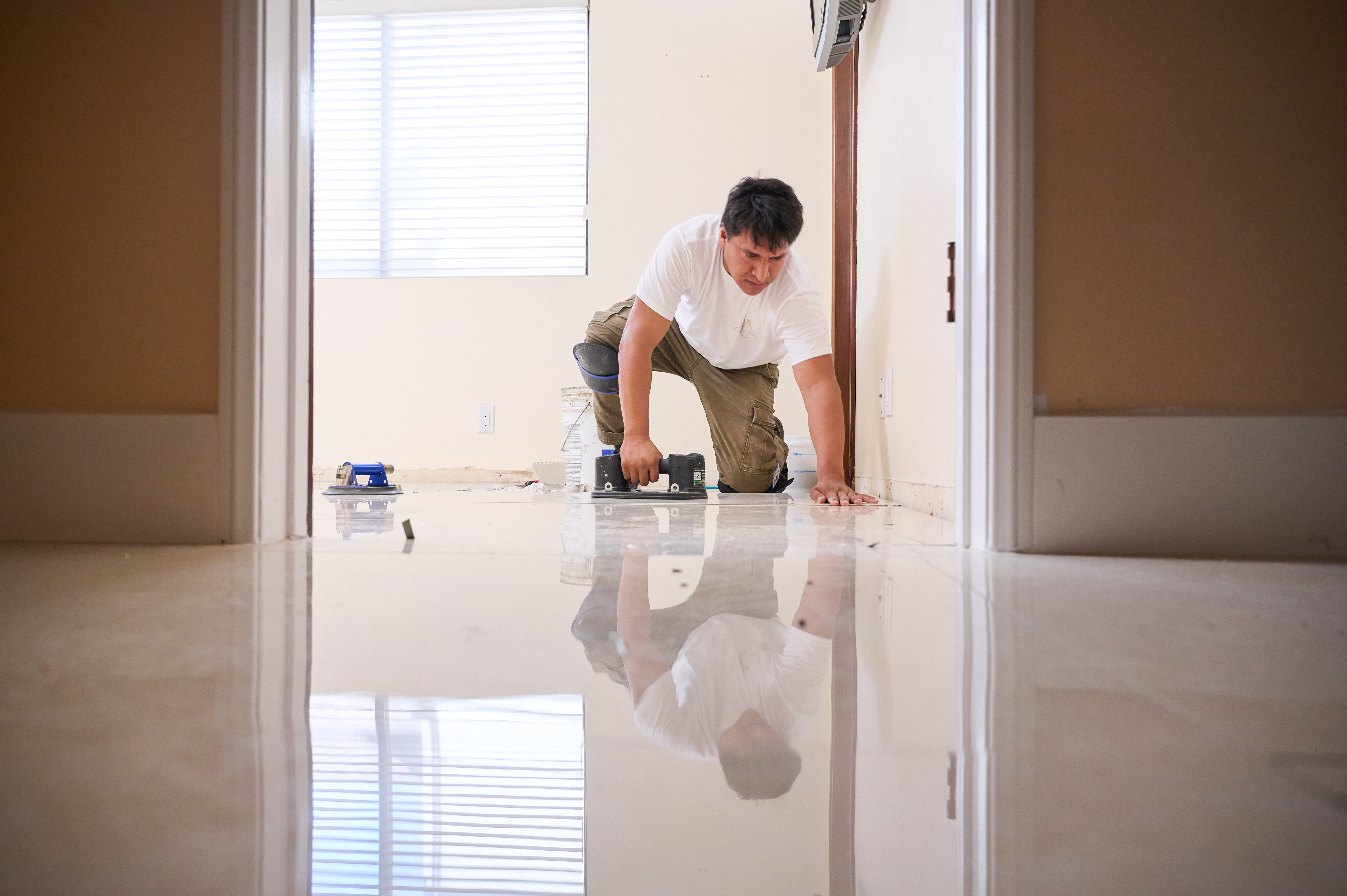 A worker in knee pads aligns a tile on the floor during installation, with his reflection visible on the polished surface.