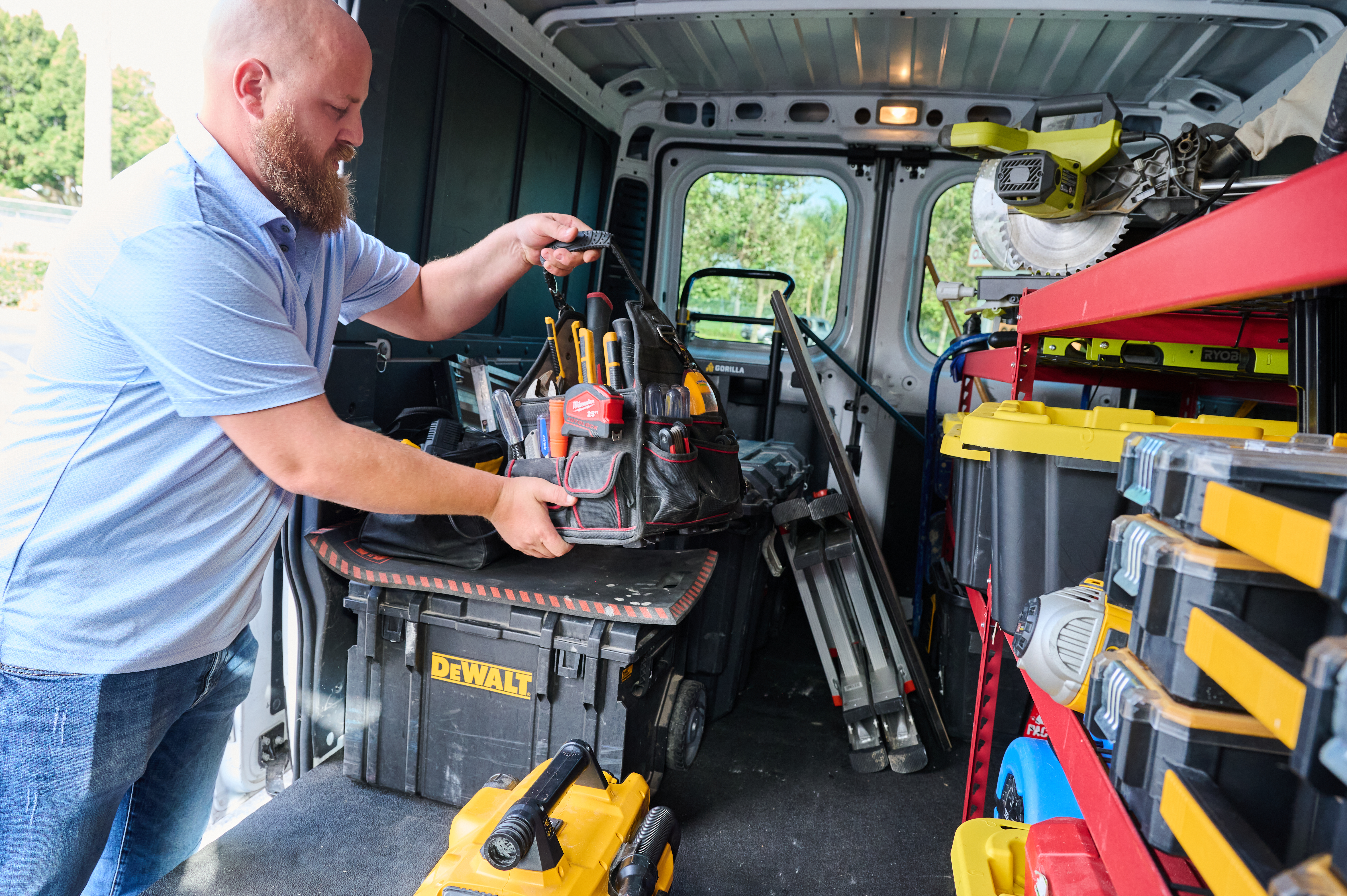 Handyman Chris McNeill from C.A. Shamrock LLC  organizing his tool bag in the back of a work van filled with equipment, toolboxes, and various tools.