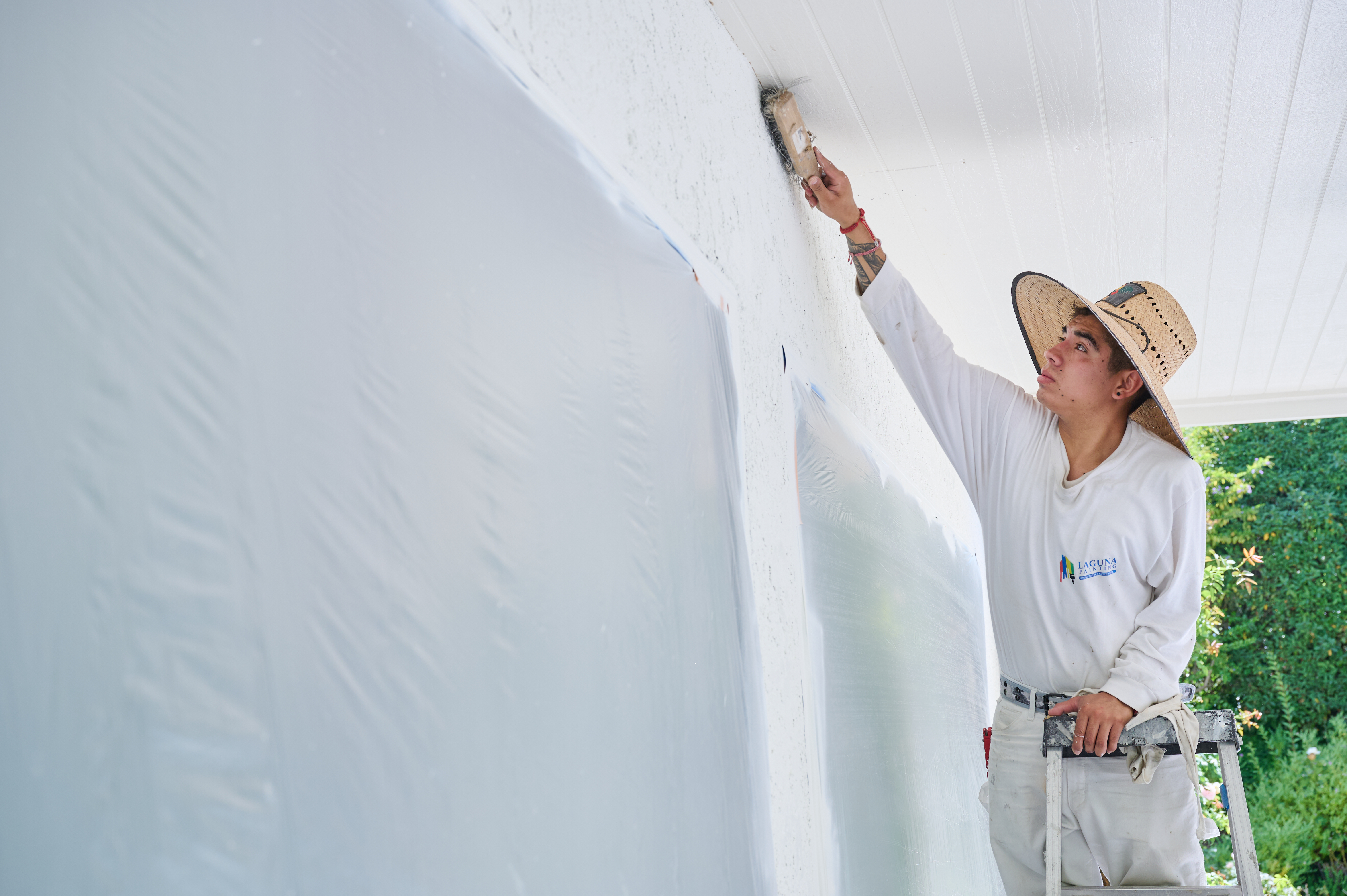 Painter brushes an upper wall corner while standing on a ladder, wearing a straw hat for sun protection.