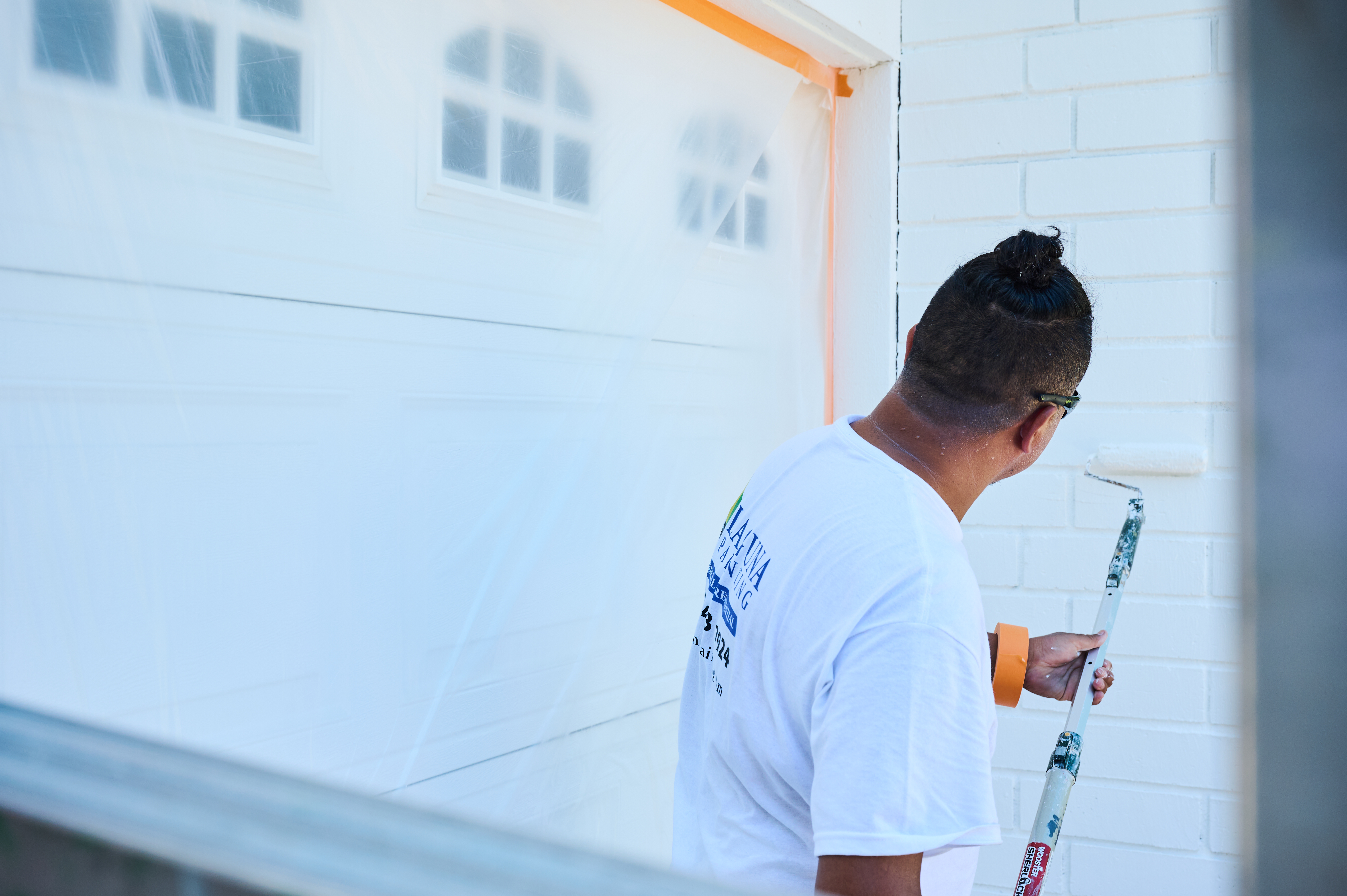 Silvio, a painter with Laguna Painting, uses a roller to paint a brick wall white, with a garage door protected in plastic.