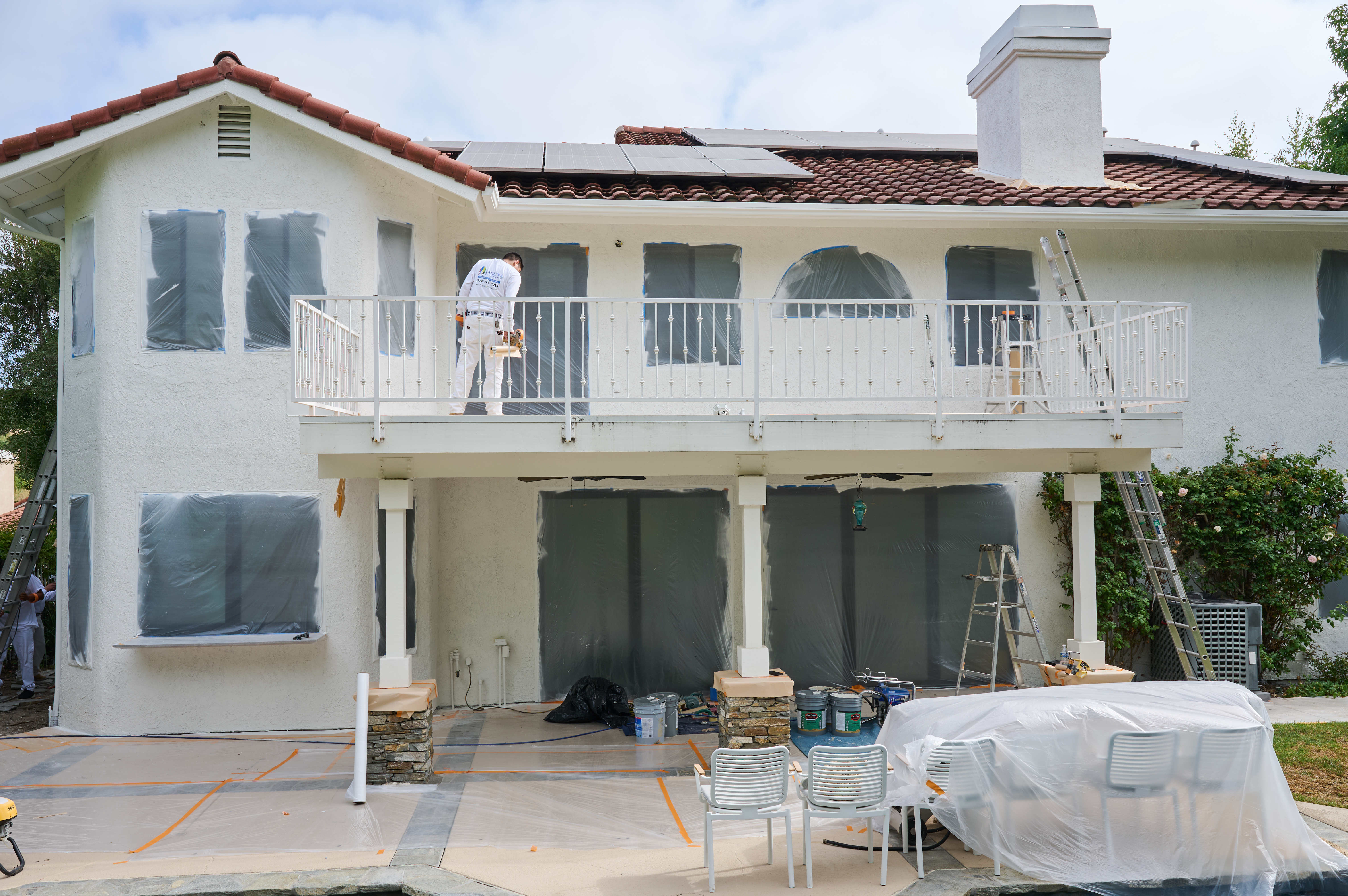 Home exterior prepared for painting with windows and furniture covered as a painter works on the balcony