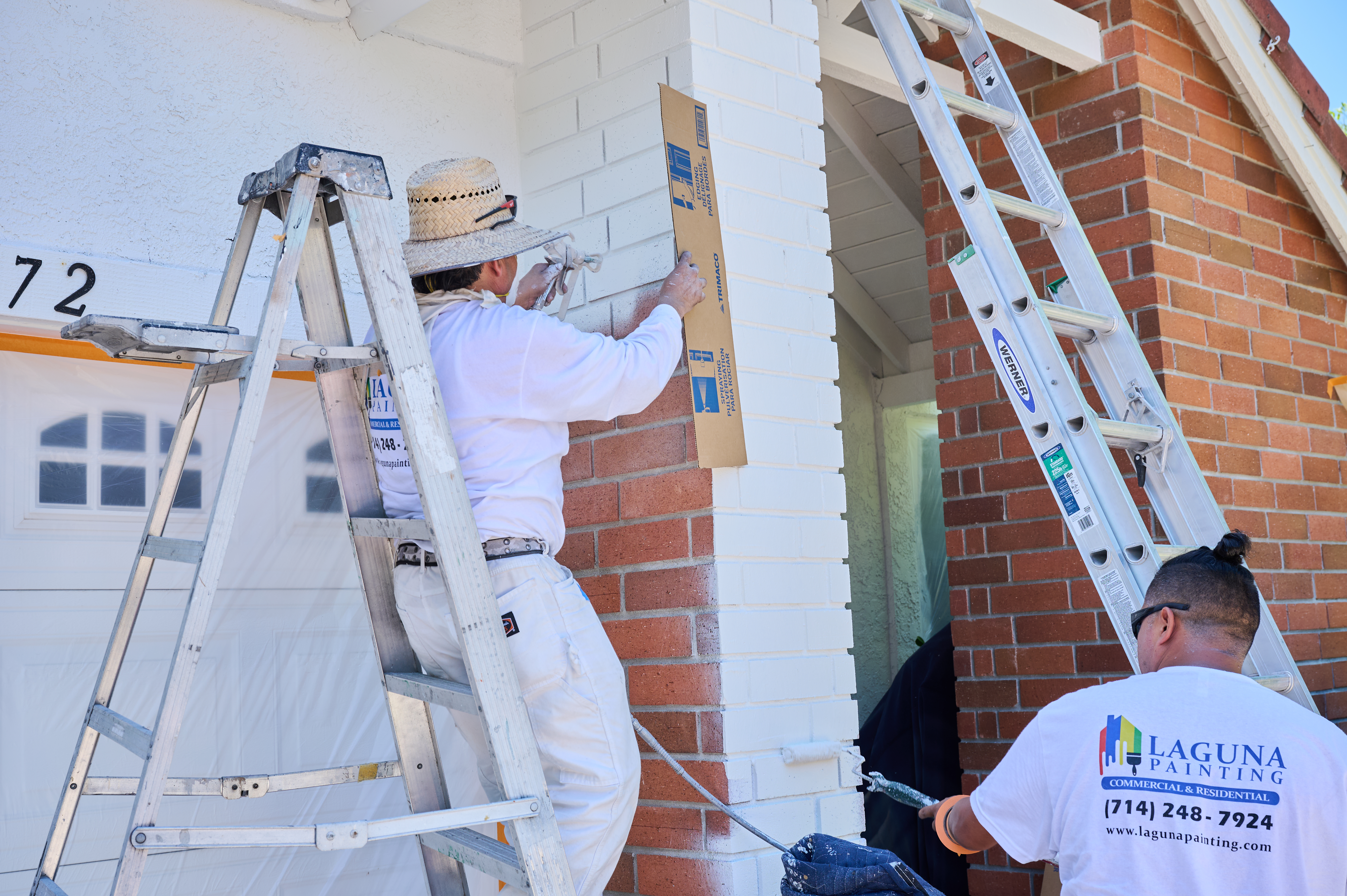 Mario and Silvio from Laguna Painting use ladders to spray paint a brick wall near a garage, carefully protecting surrounding areas.