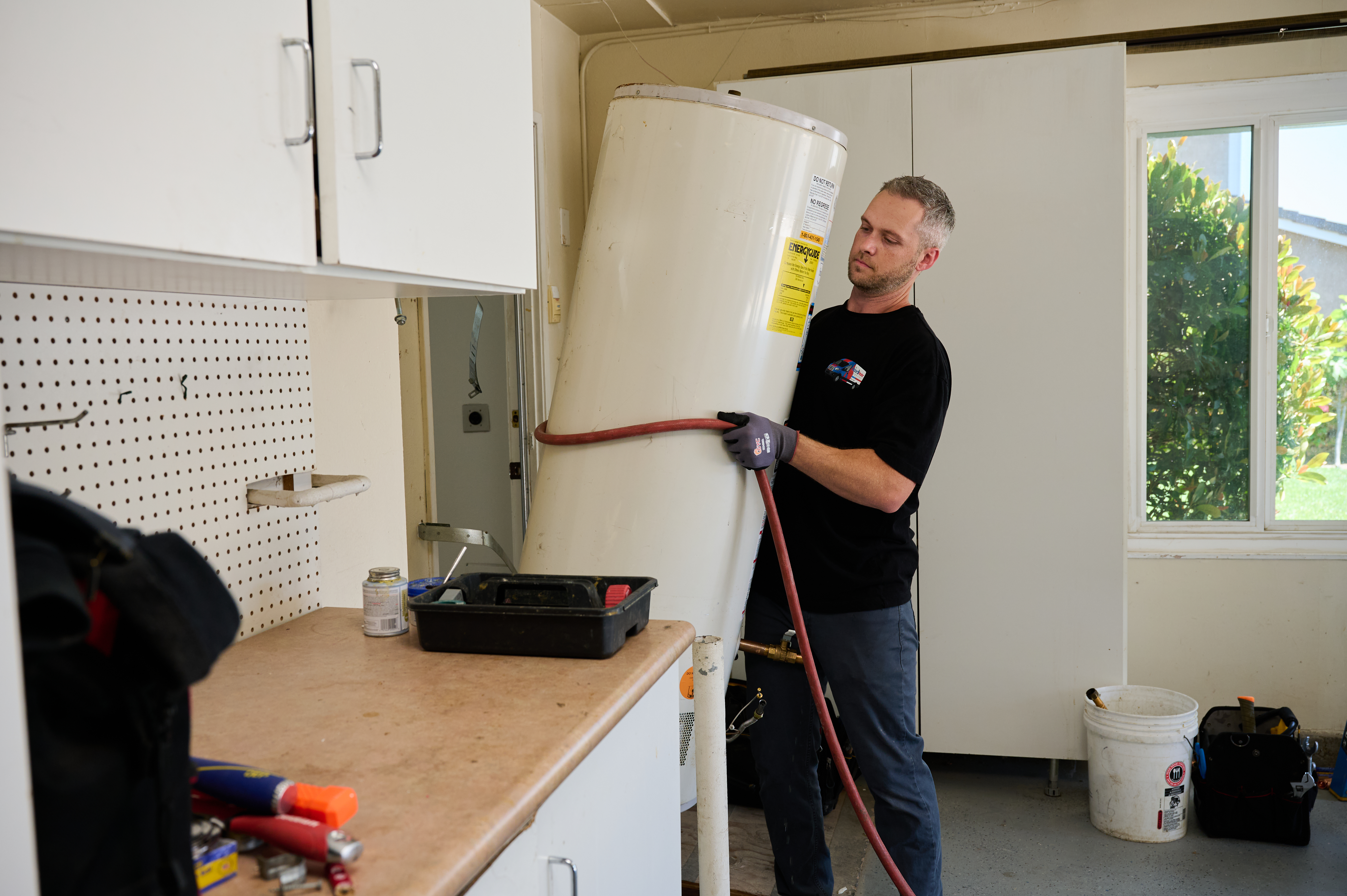 Technician carefully lifts a water heater while wearing gloves in a well-lit garage with tools and supplies on the counter nearby.