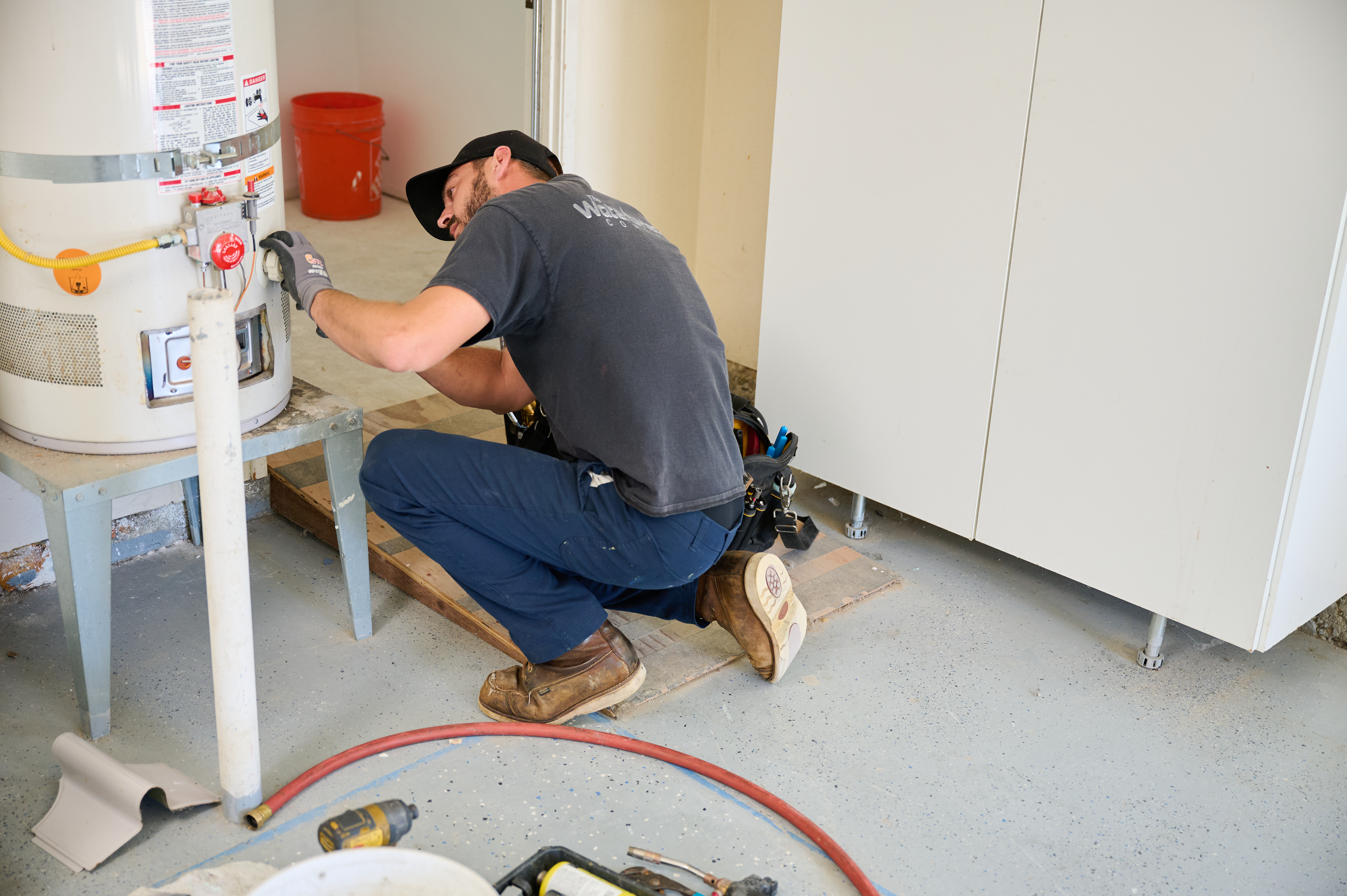 A technician kneels on the floor to inspect a water heater in a garage, surrounded by tools, hoses, and equipment.
