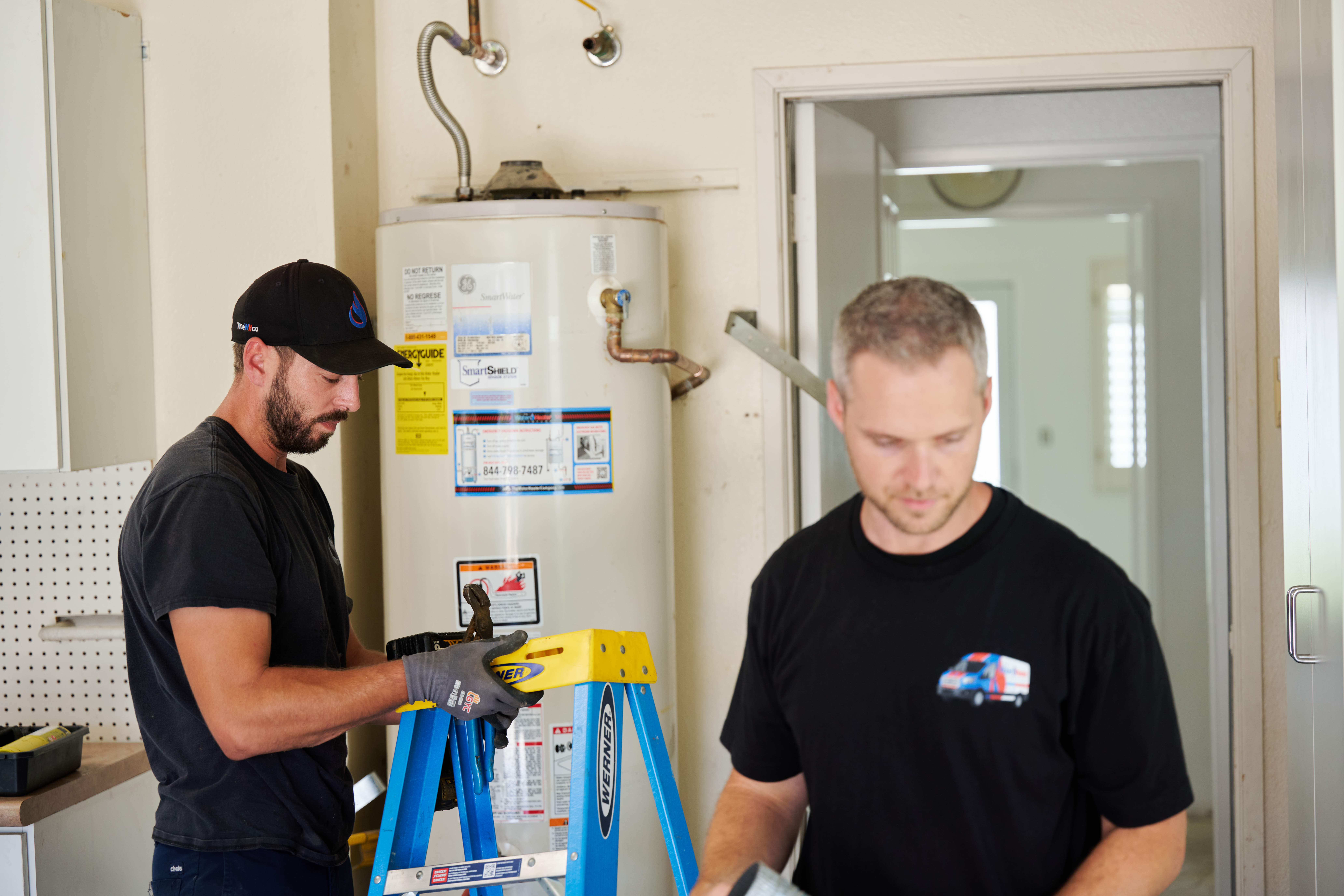 Two technicians work near a water heater; one adjusts tools on a ladder, while the other is focused on equipment in the foreground.