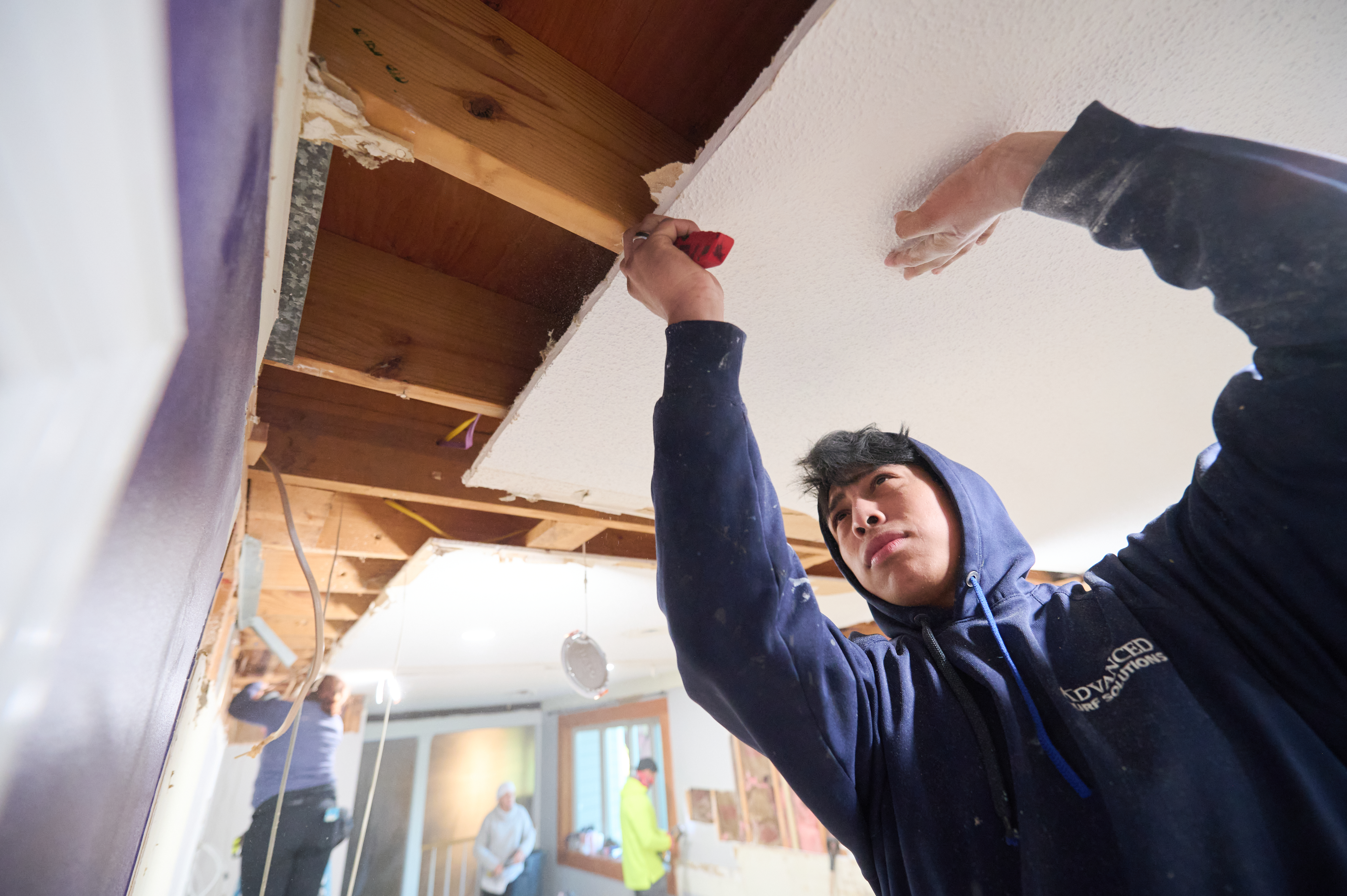 A worker in a blue hoodie carefully removes a ceiling panel to expose wooden beams, with other team members visible in the background.