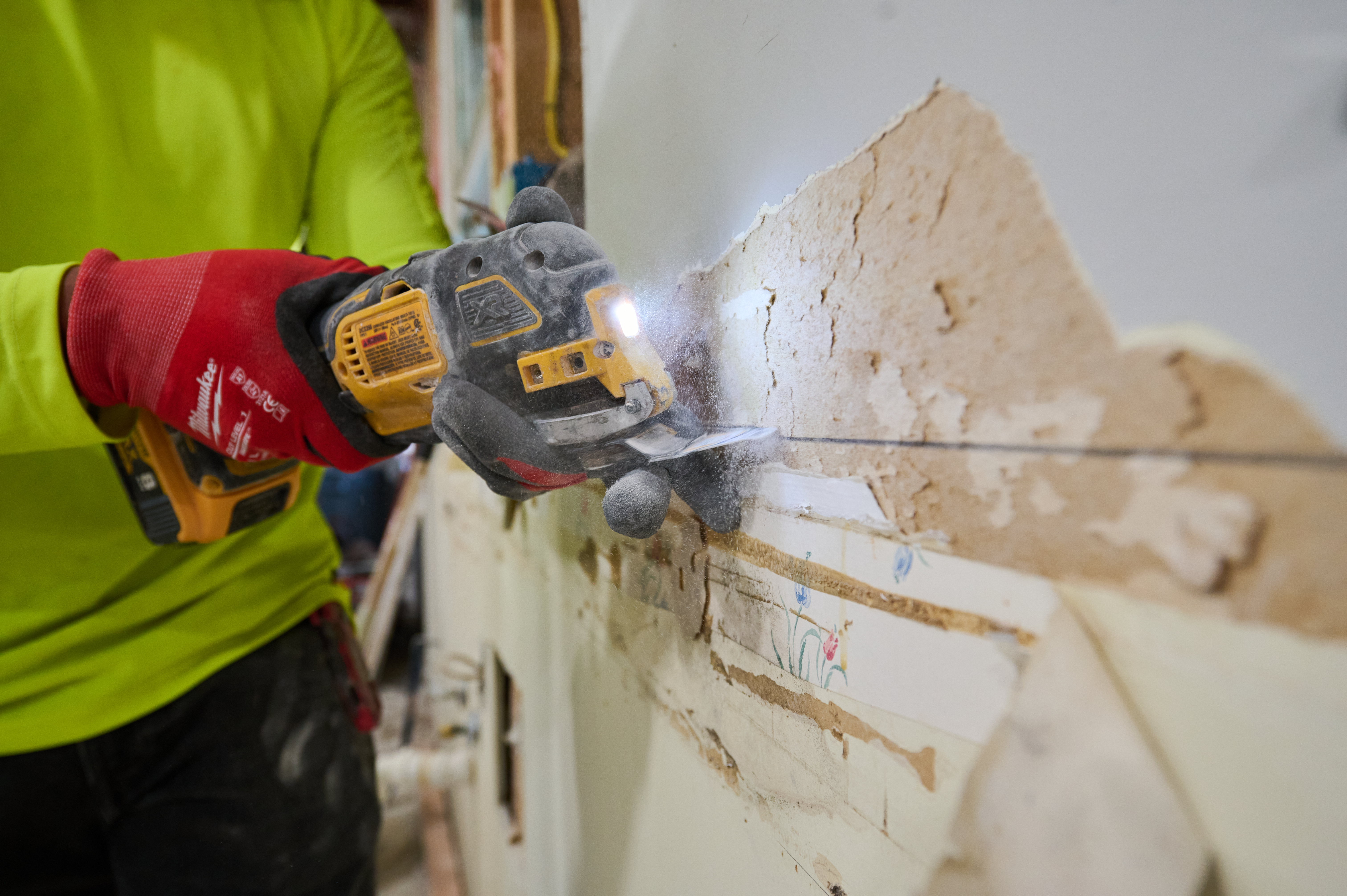 Close-up of a power tool cutting through drywall, with red-gloved hands holding the tool as dust and debris scatter.