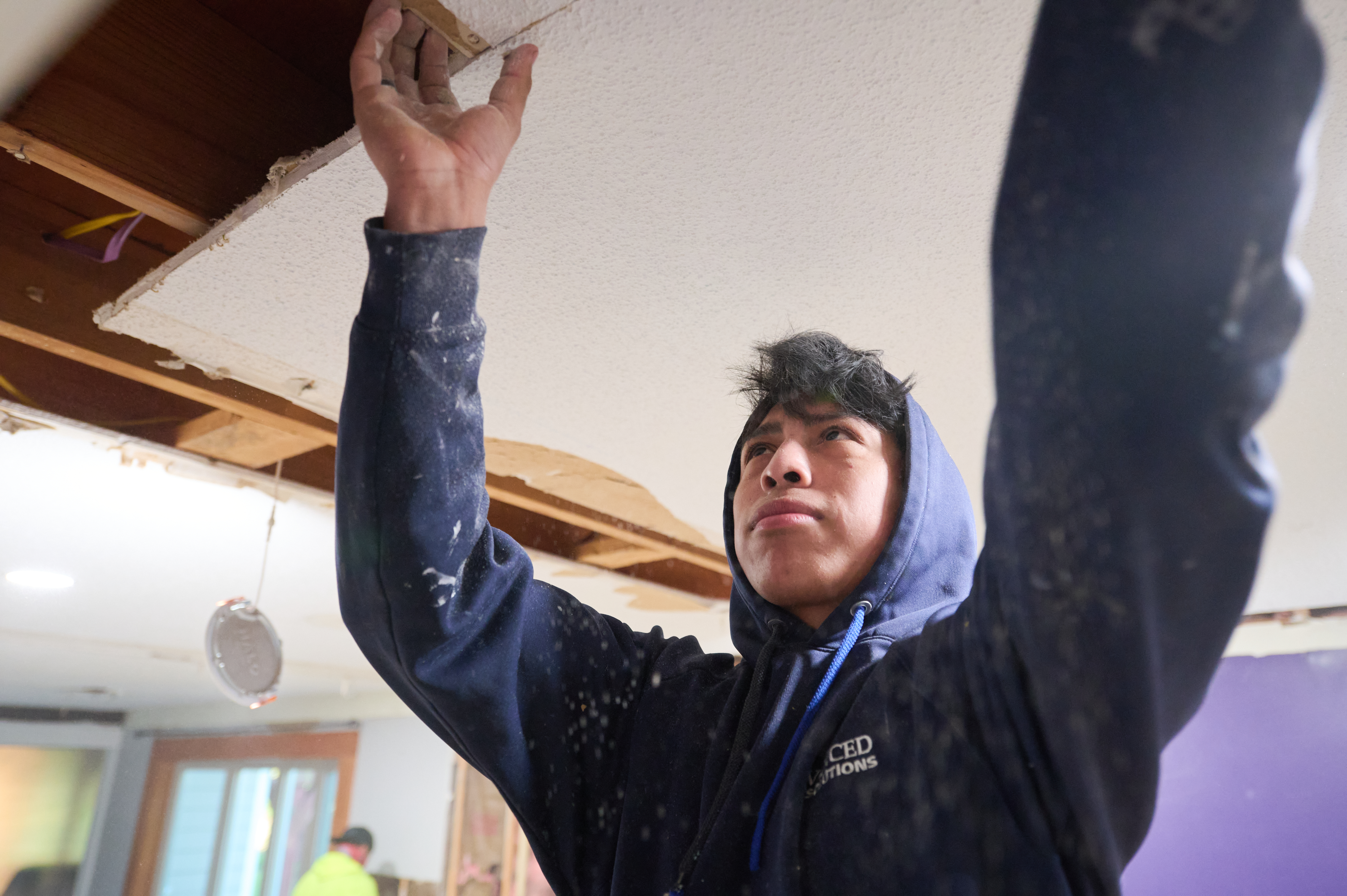 A worker in a blue hoodie carefully removing a section of the ceiling during a renovation project, exposing wooden beams above.