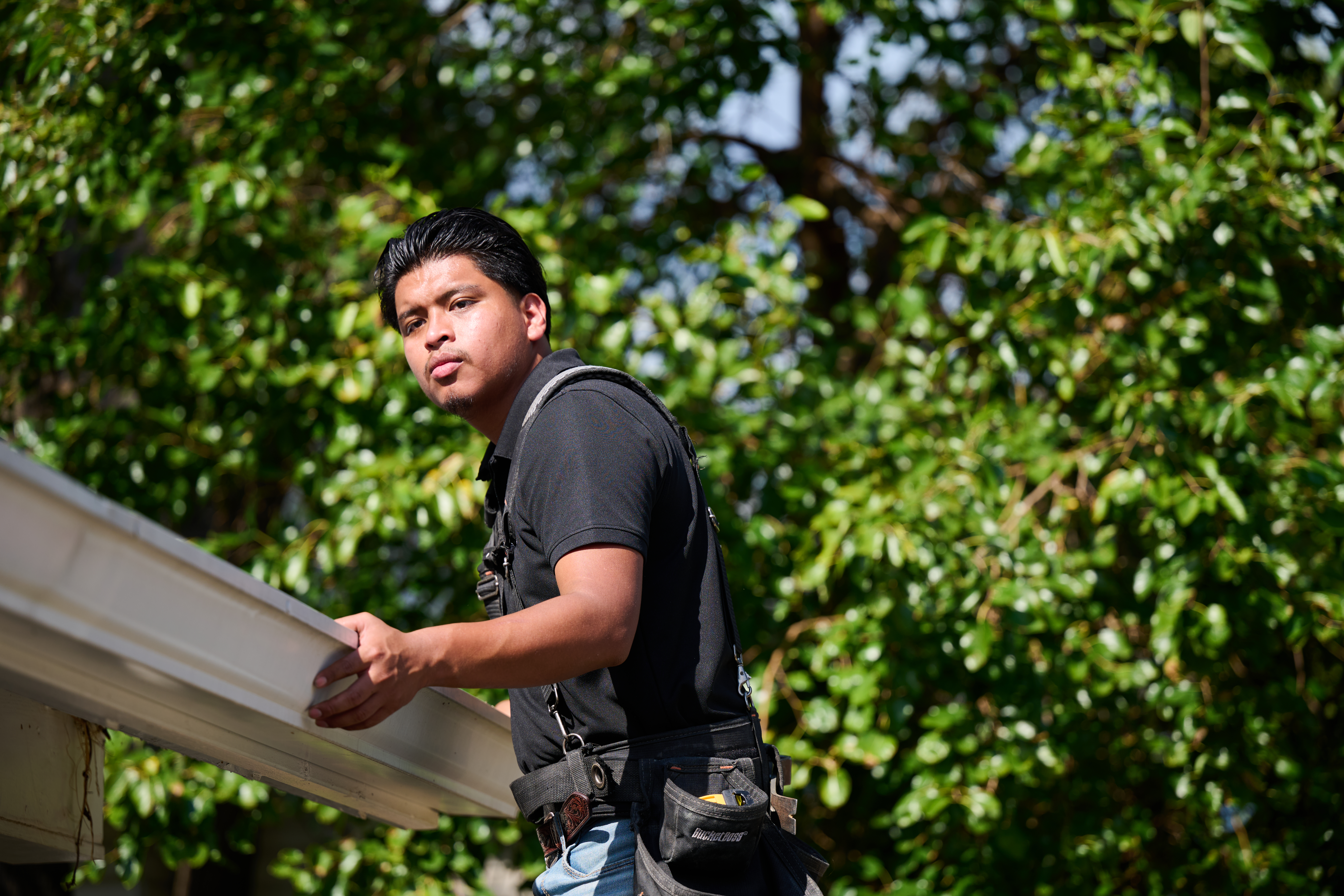 Worker in a tool harness inspects a gutter under bright sunlight, surrounded by green foliage in the background.