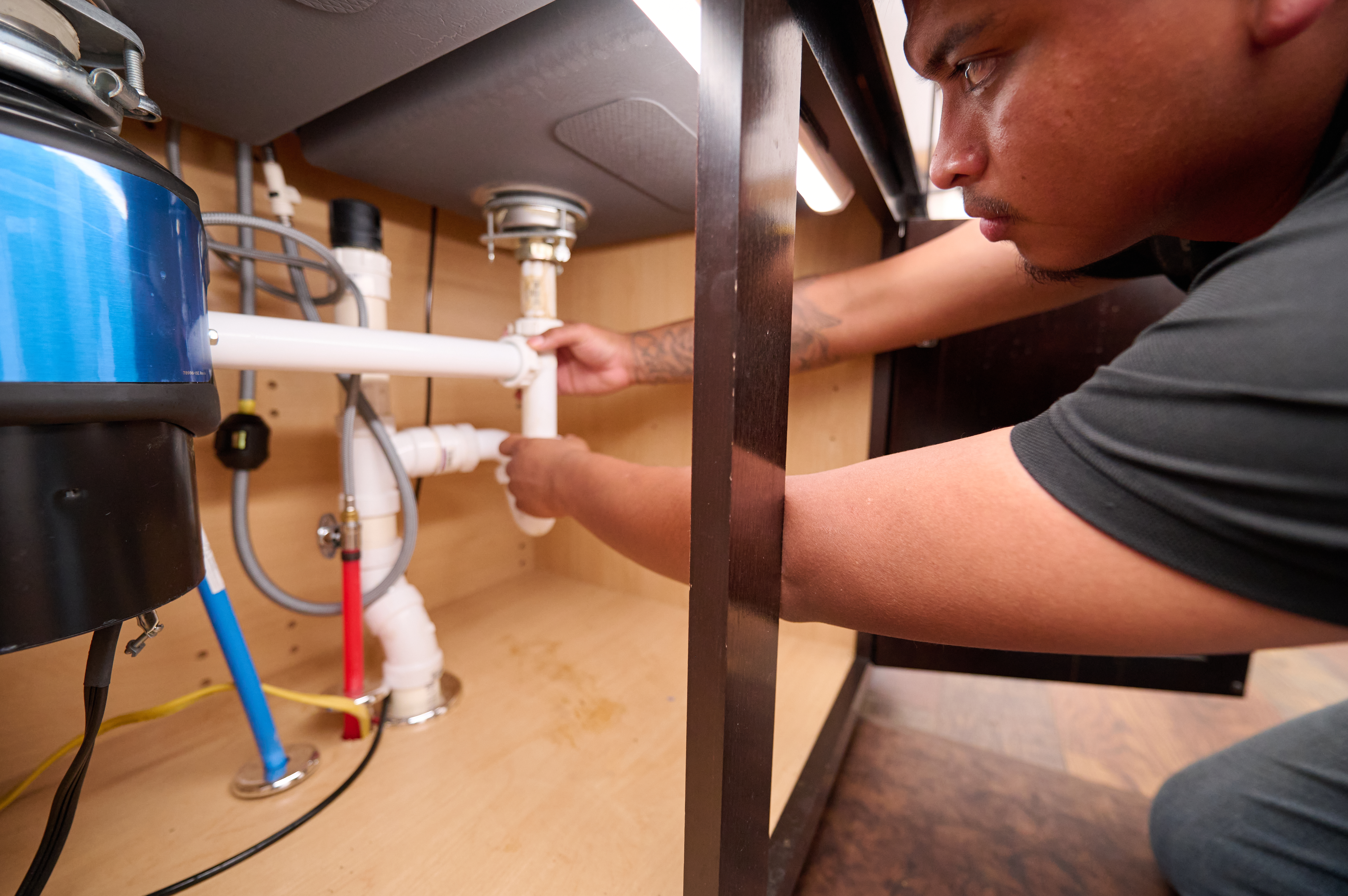 Plumber installing PVC pipes under a kitchen sink, connecting drainage system and garbage disposal in a cabinet space.
