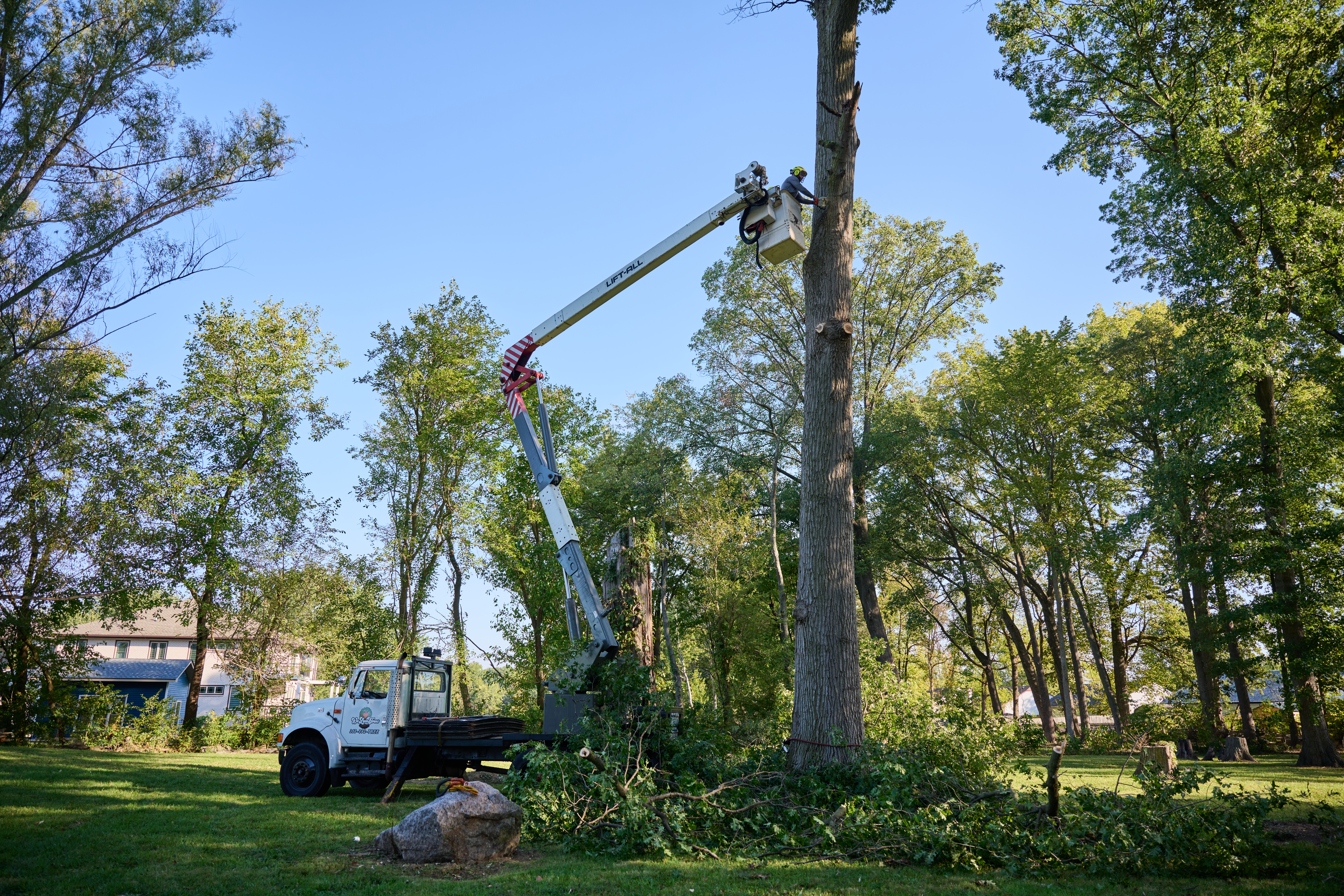 White flatbed truck with company logo is parked on grass near a large boulder and tree debris in a residential area.