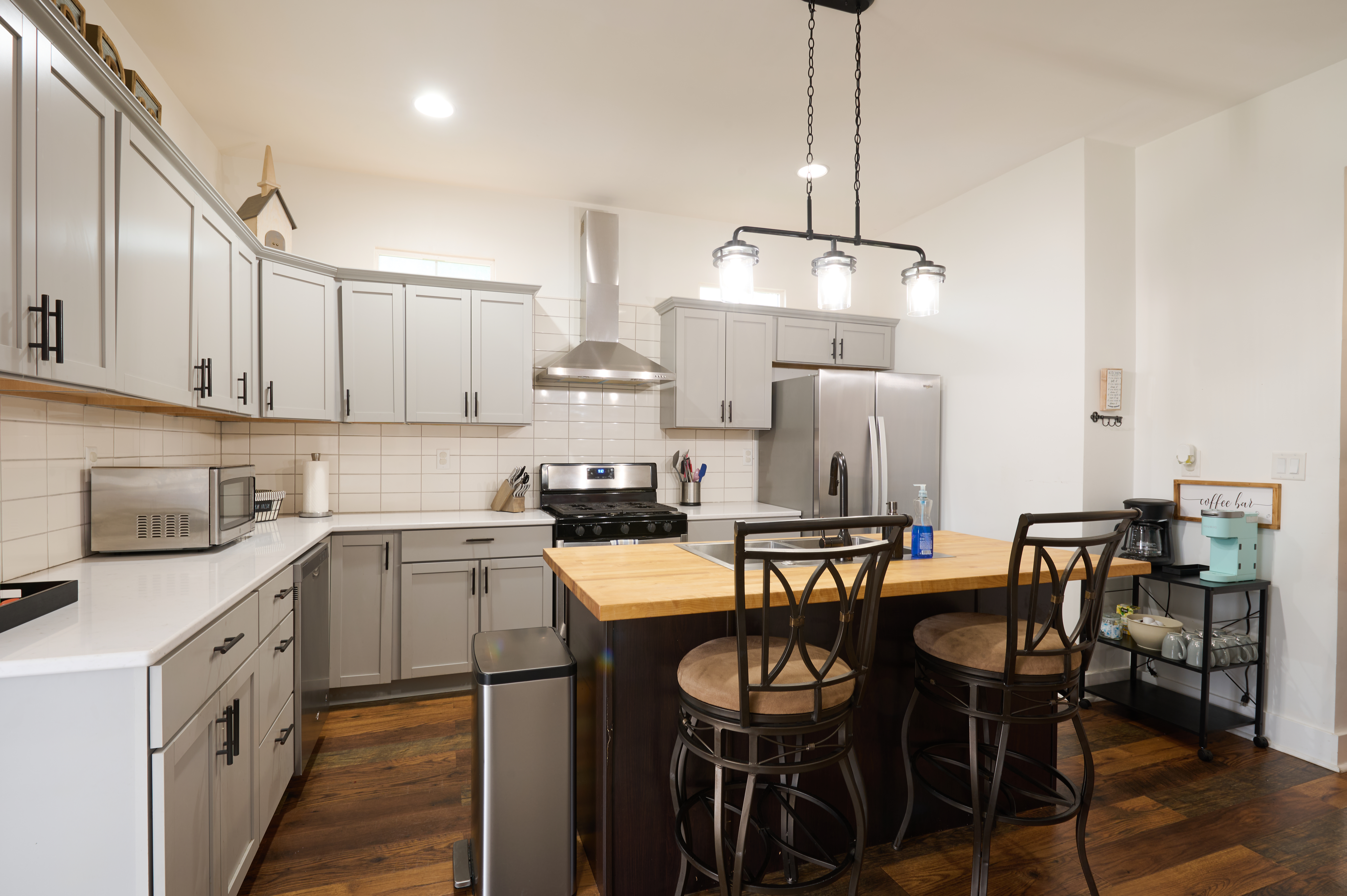 A modern kitchen with gray cabinets, a center island with wooden countertop and bar stools, stainless steel appliances, and pendant lighting.