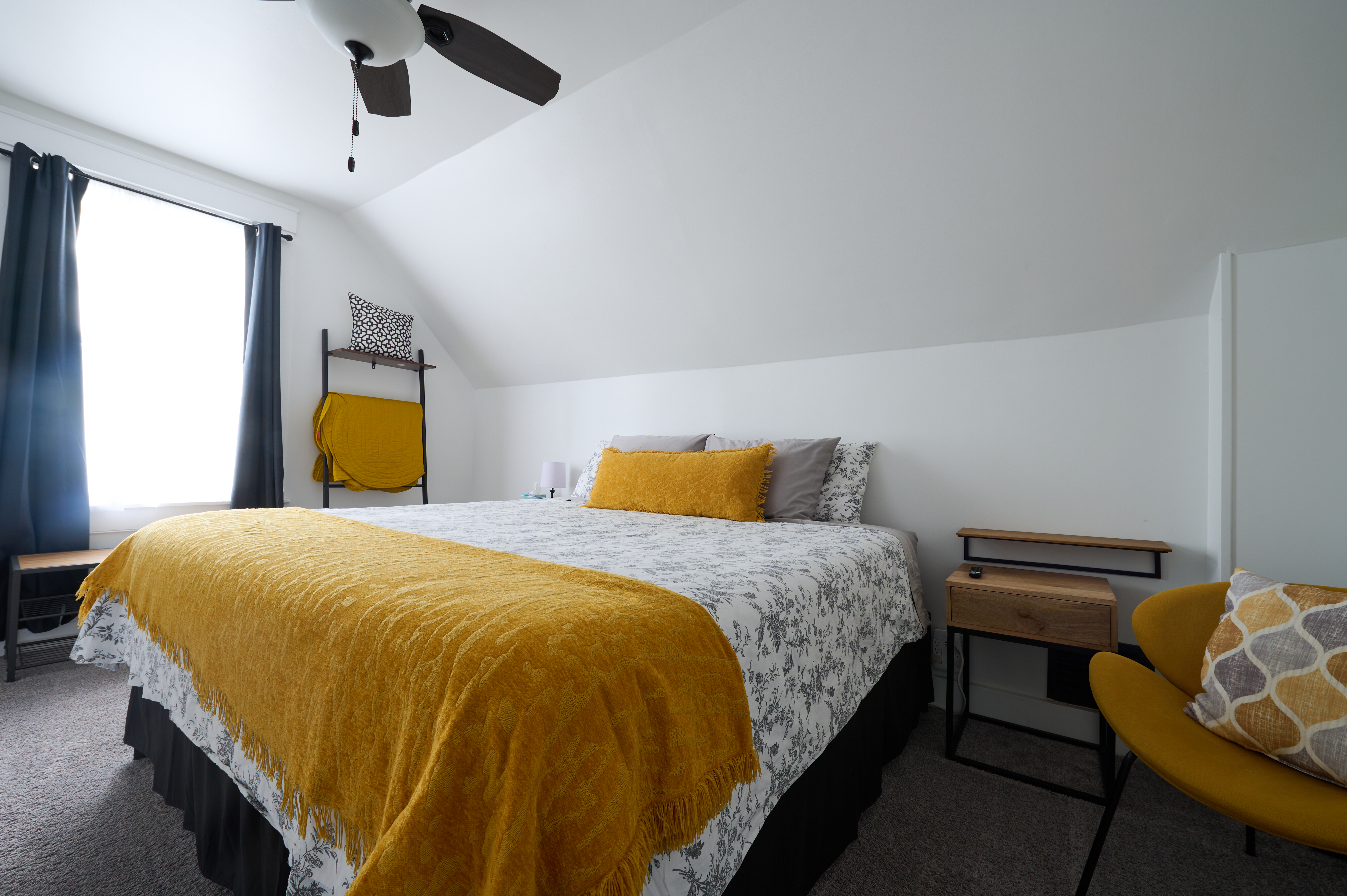 A cozy bedroom with a sloped ceiling, featuring a bed with floral bedding and a mustard yellow throw, and modern furnishings.