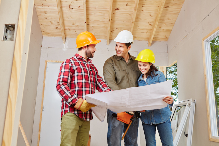 three adults in hard hats smile and look over renovation plans in a partially finished room