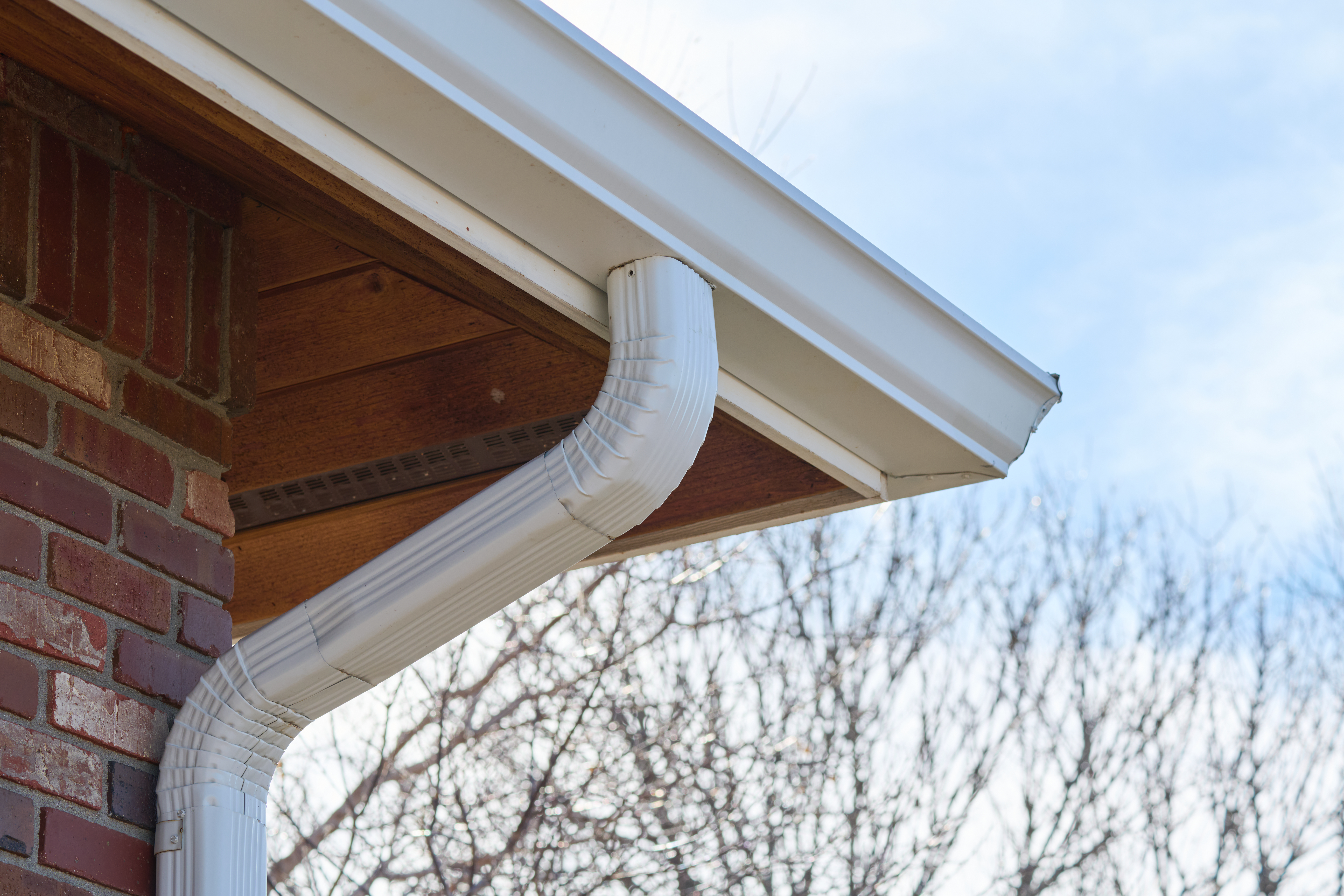 White downspout connected to seamless gutter system on brick home, directing rainwater away from the roof structure.