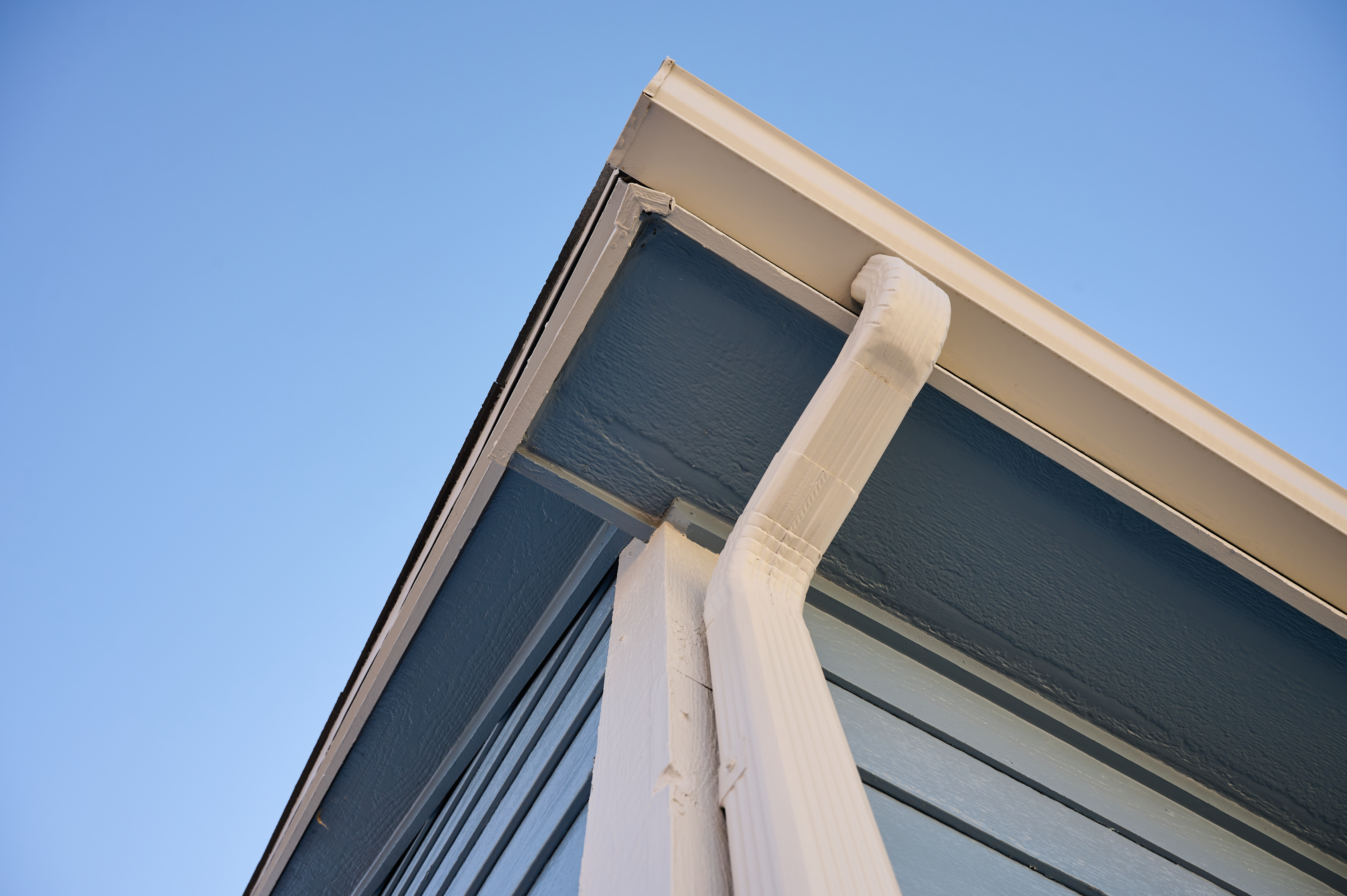 White downspout connected to seamless gutter on a blue-painted home exterior, channeling rainwater away effectively.