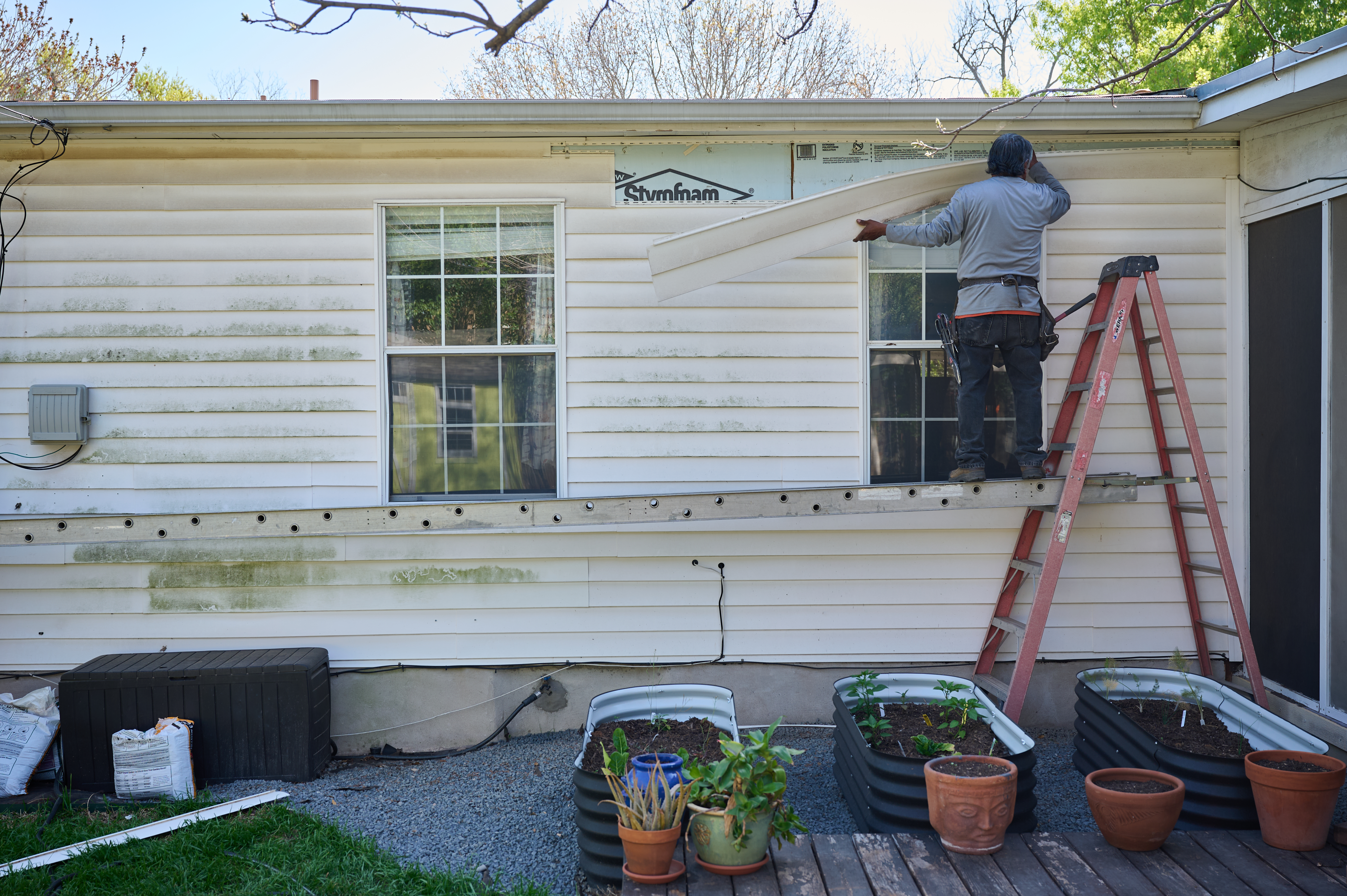 Worker replacing exterior vinyl siding on a residential home using a ladder and scaffold, with garden planters below