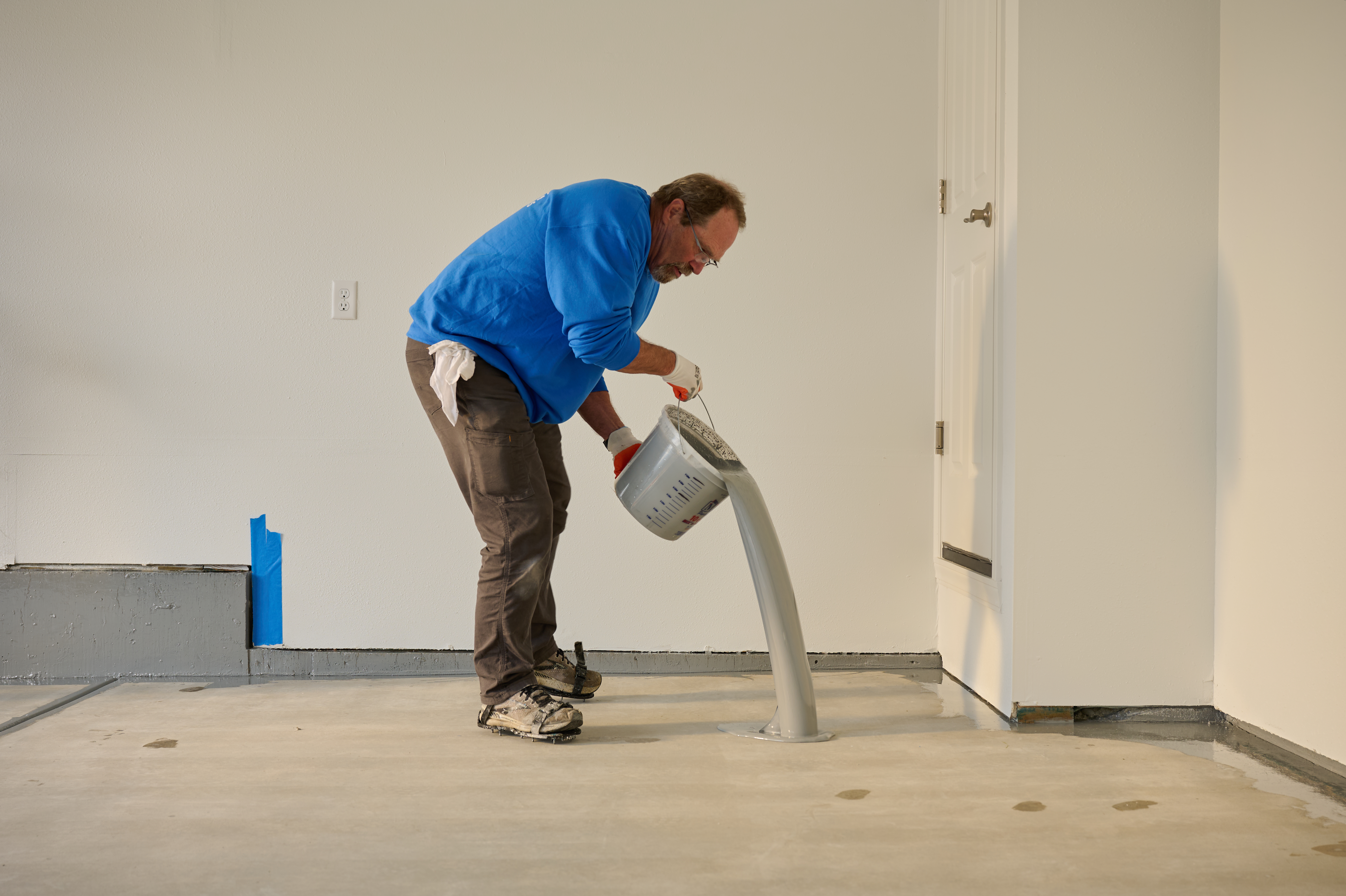 Worker pouring gray epoxy coating onto a concrete floor, preparing the surface for a smooth, durable finish.