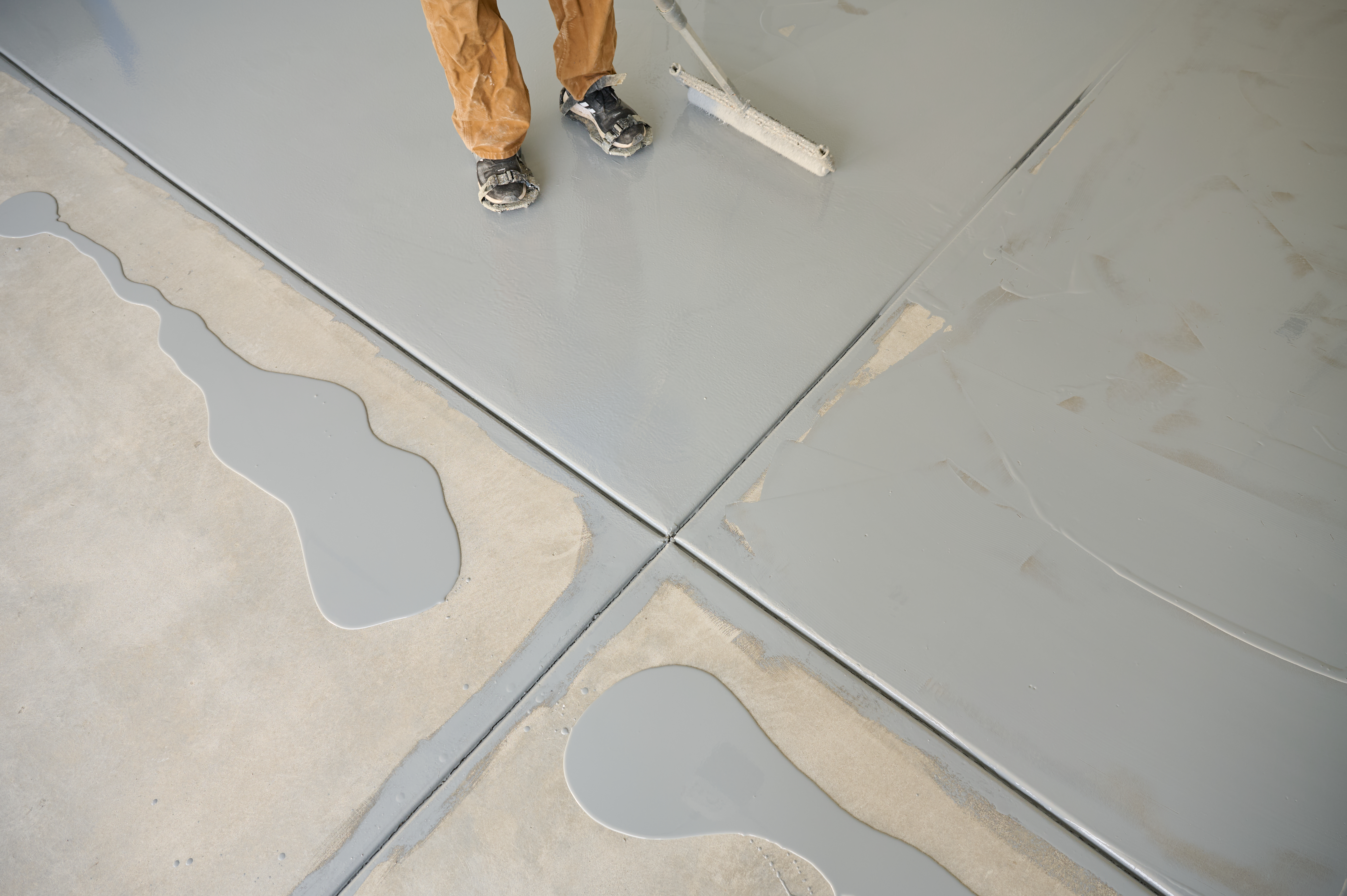 Worker applying gray epoxy coating to concrete floor, with patches of wet epoxy and visible roller tool in use