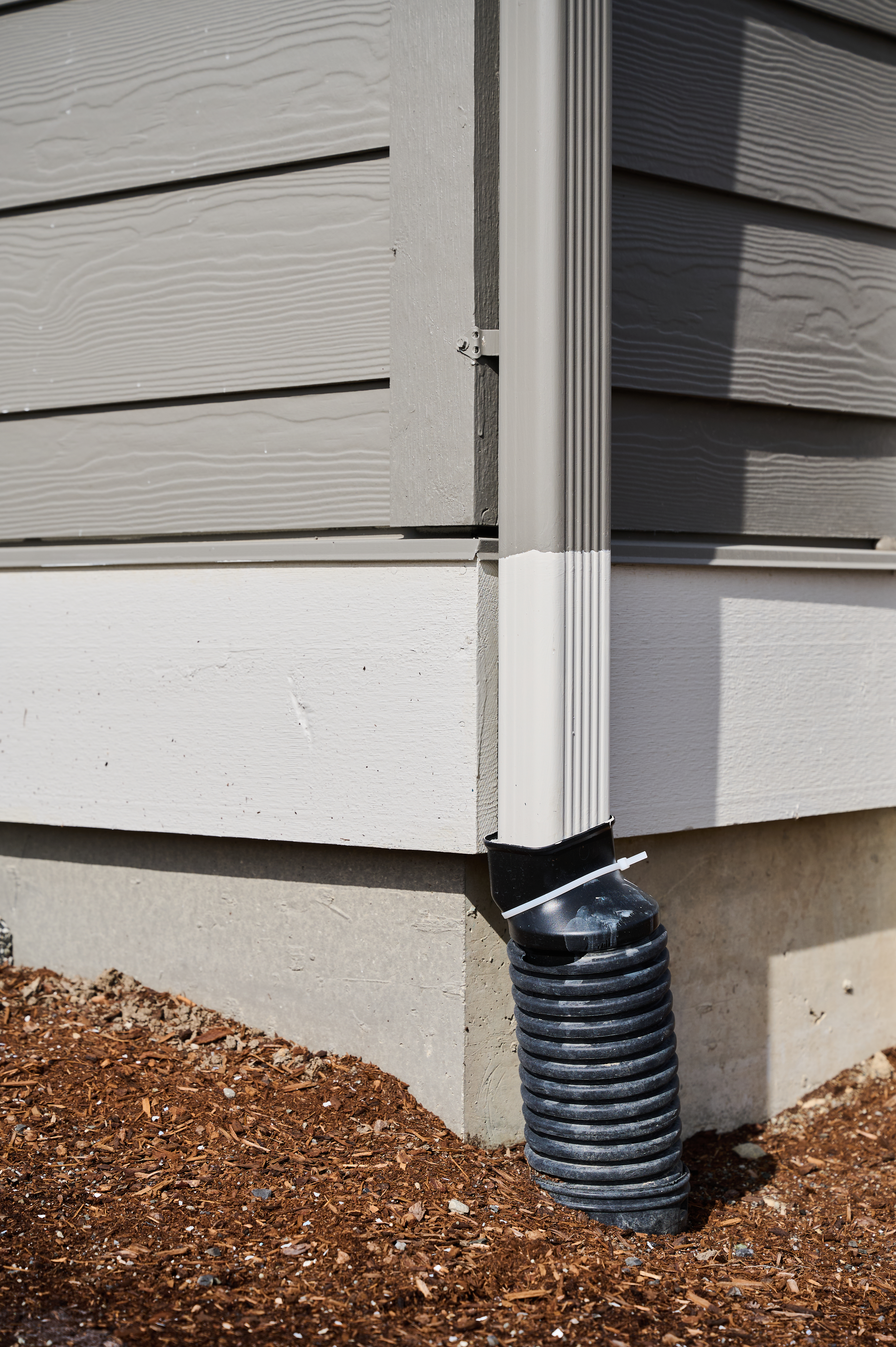 Flexible downspout extension secured with a zip tie, directing water away from a home's foundation onto mulch ground.
