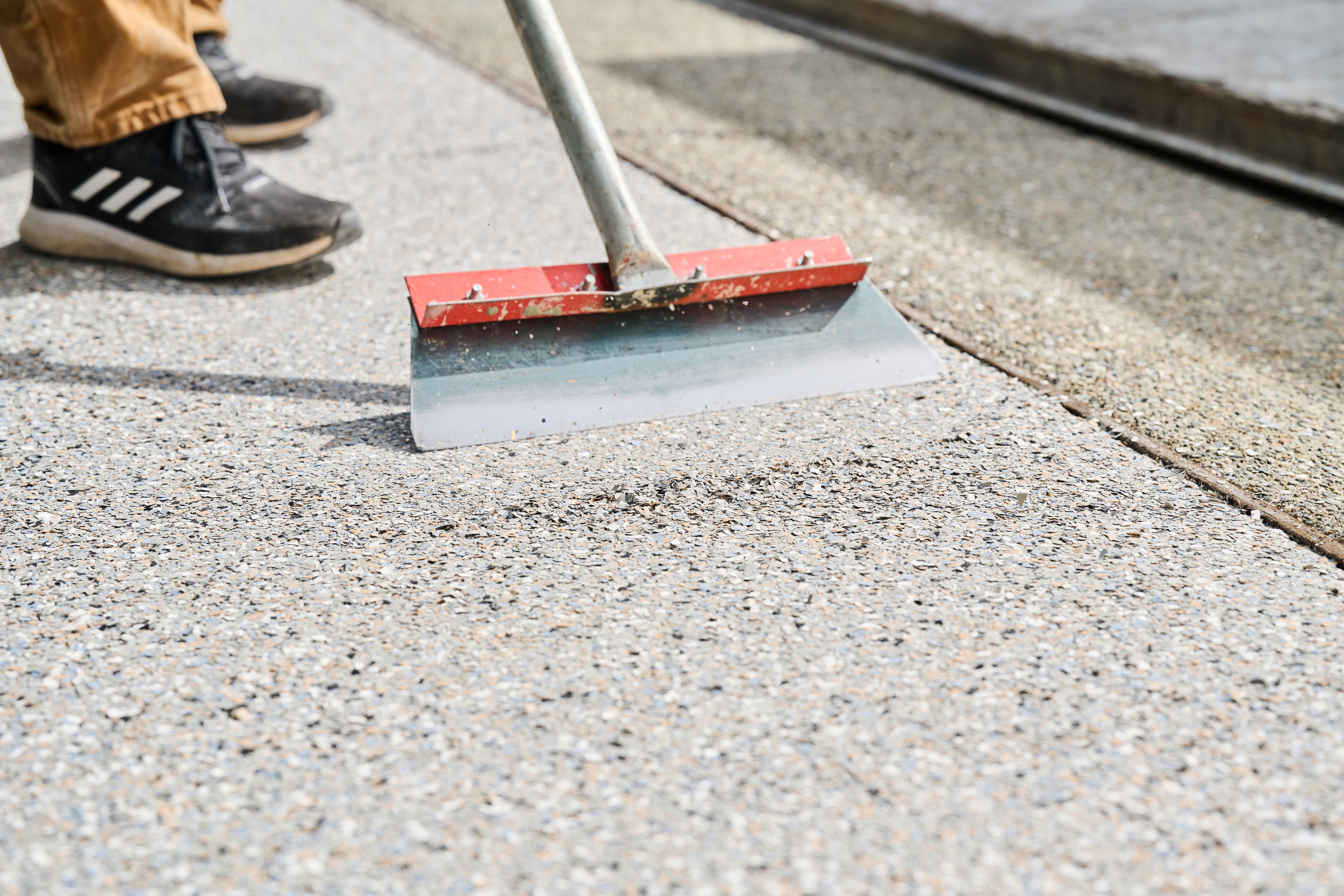 Worker using a red-edged floor scraper to smooth and level concrete surface with a textured, pebble-finish coating