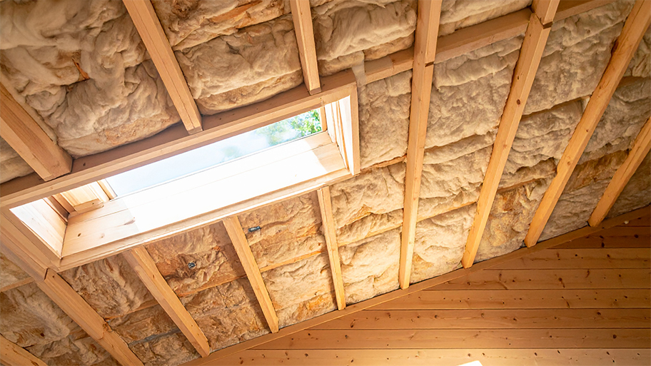 An unfinished attic ceiling shows exposed wooden rafters filled with thick insulation around a skylight
