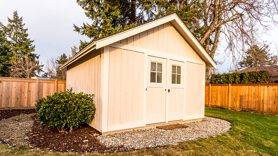 Beige backyard storage shed with double doors, surrounded by mulch, gravel, and a wooden fence