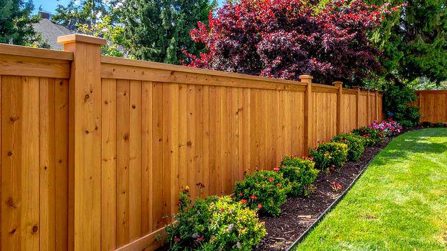 A wooden privacy fence with decorative posts borders a manicured lawn and a landscaped garden bed